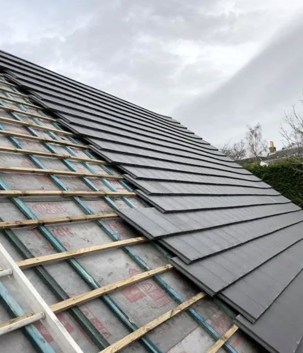Two-story beige house with dark trim and shutters, on a grassy hill, under a clear blue sky.