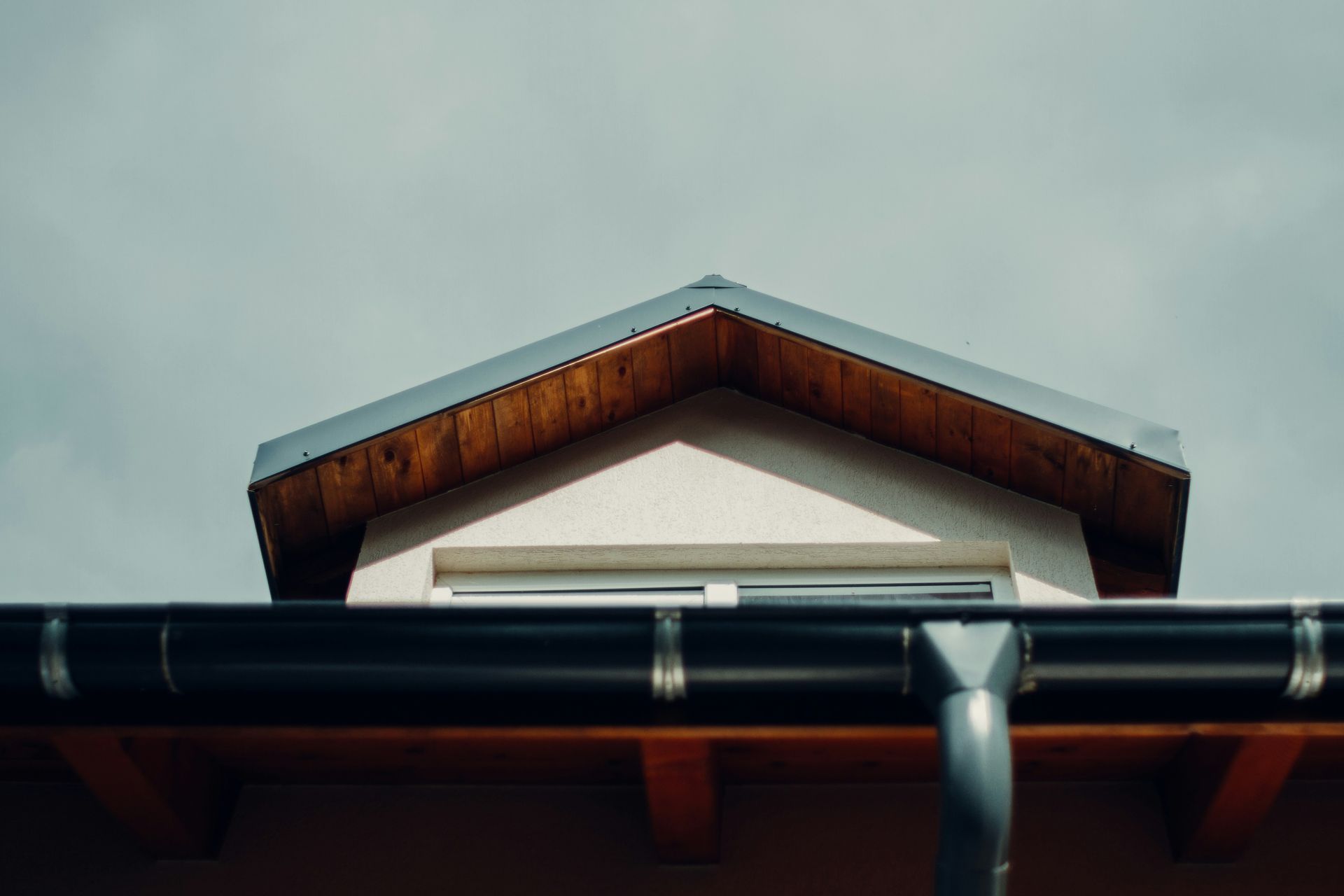 Dormer with black roof and gutters, wooden trim, and a white stucco wall under a cloudy sky.