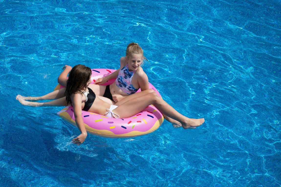 Two girls on a pink donut float in a blue swimming pool. One girl looks at the camera. Sunny day.