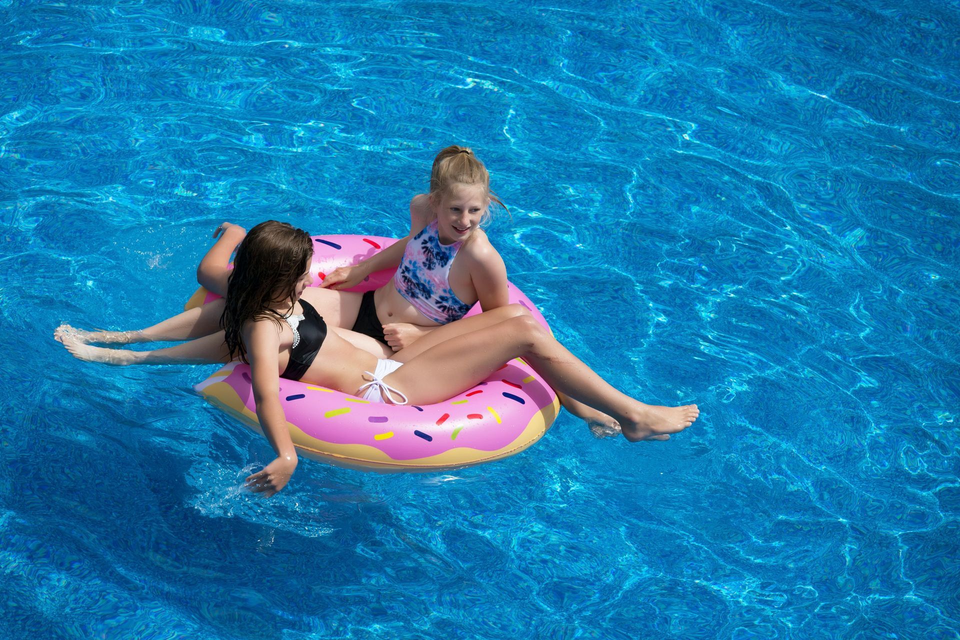 Two girls on a pink donut float in a blue swimming pool. One girl looks at the camera. Sunny day.