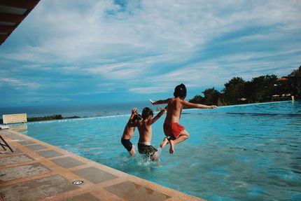 Three children jumping into a blue pool on a sunny day near the ocean.