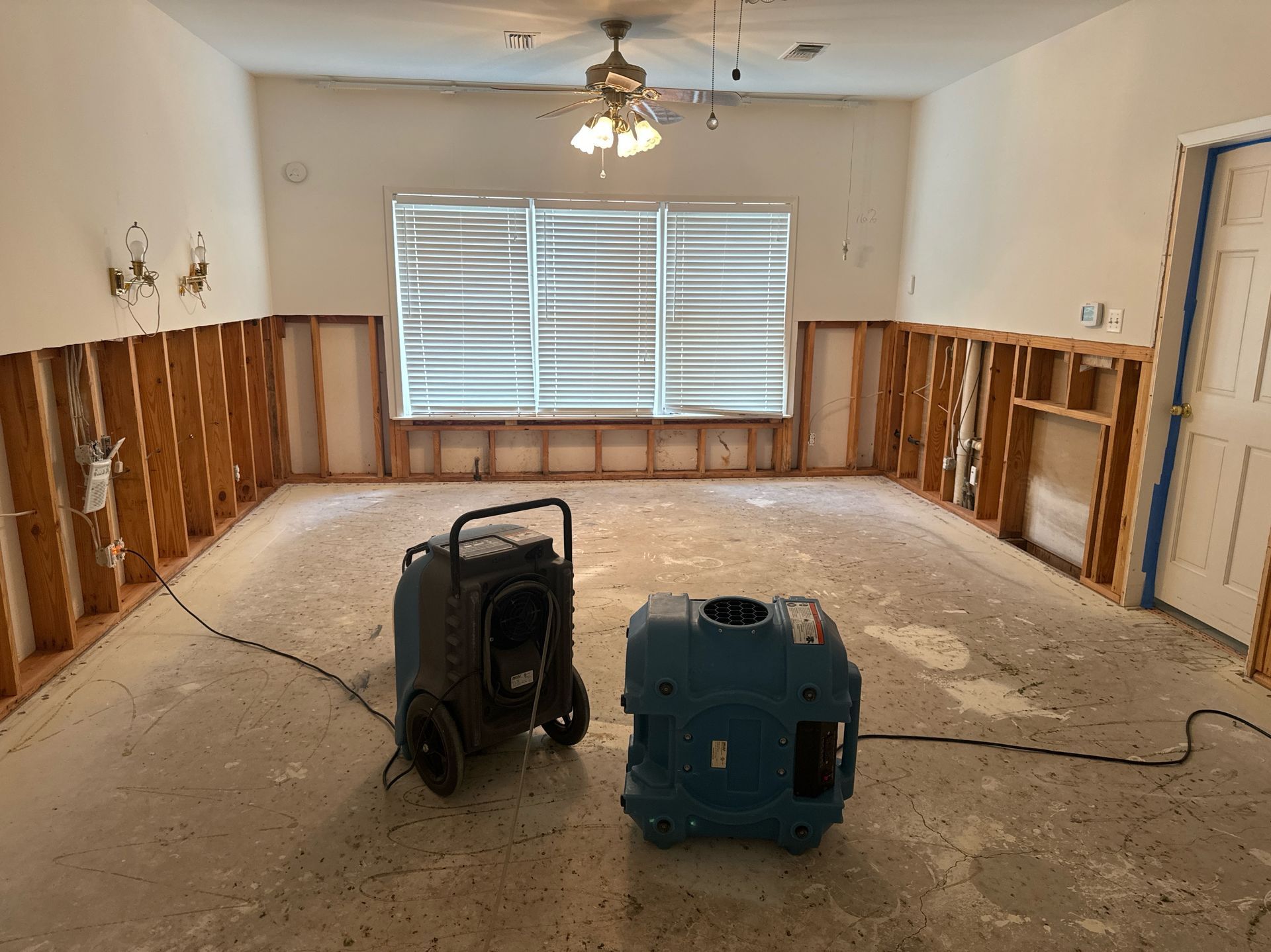 A room undergoing flood damage restoration, with exposed wall studs, two dehumidifiers, and water-damaged flooring.