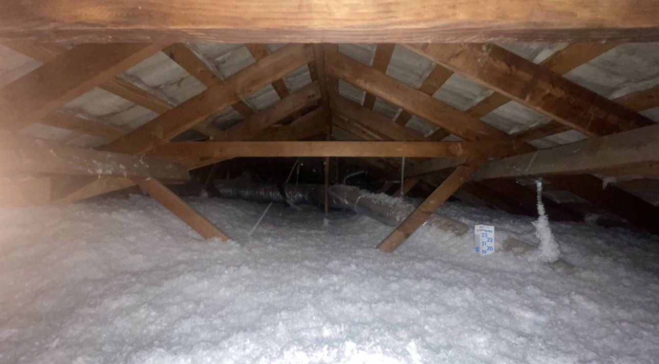 Interior view of an attic, showing wooden rafters, insulation on the floor, and a dark space in the middle.