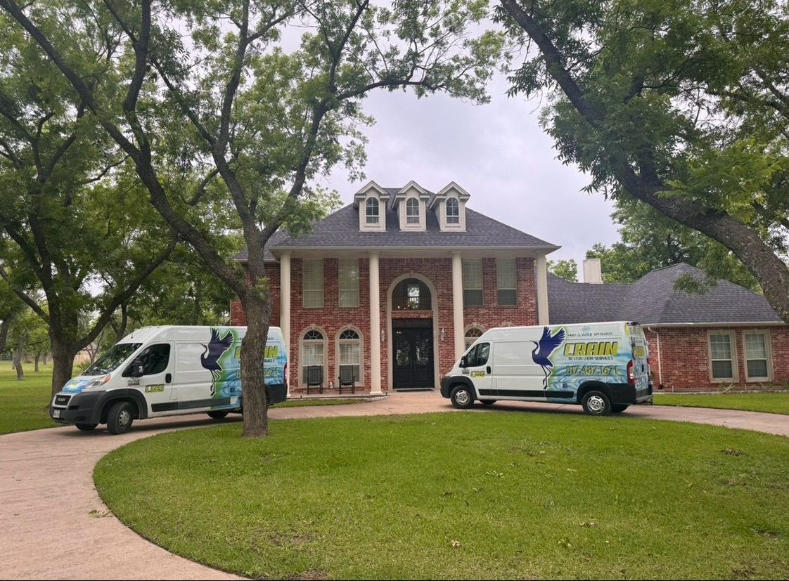 Two service vans parked in front of a large brick house, ready for work. Lush green lawn and trees frame the building.