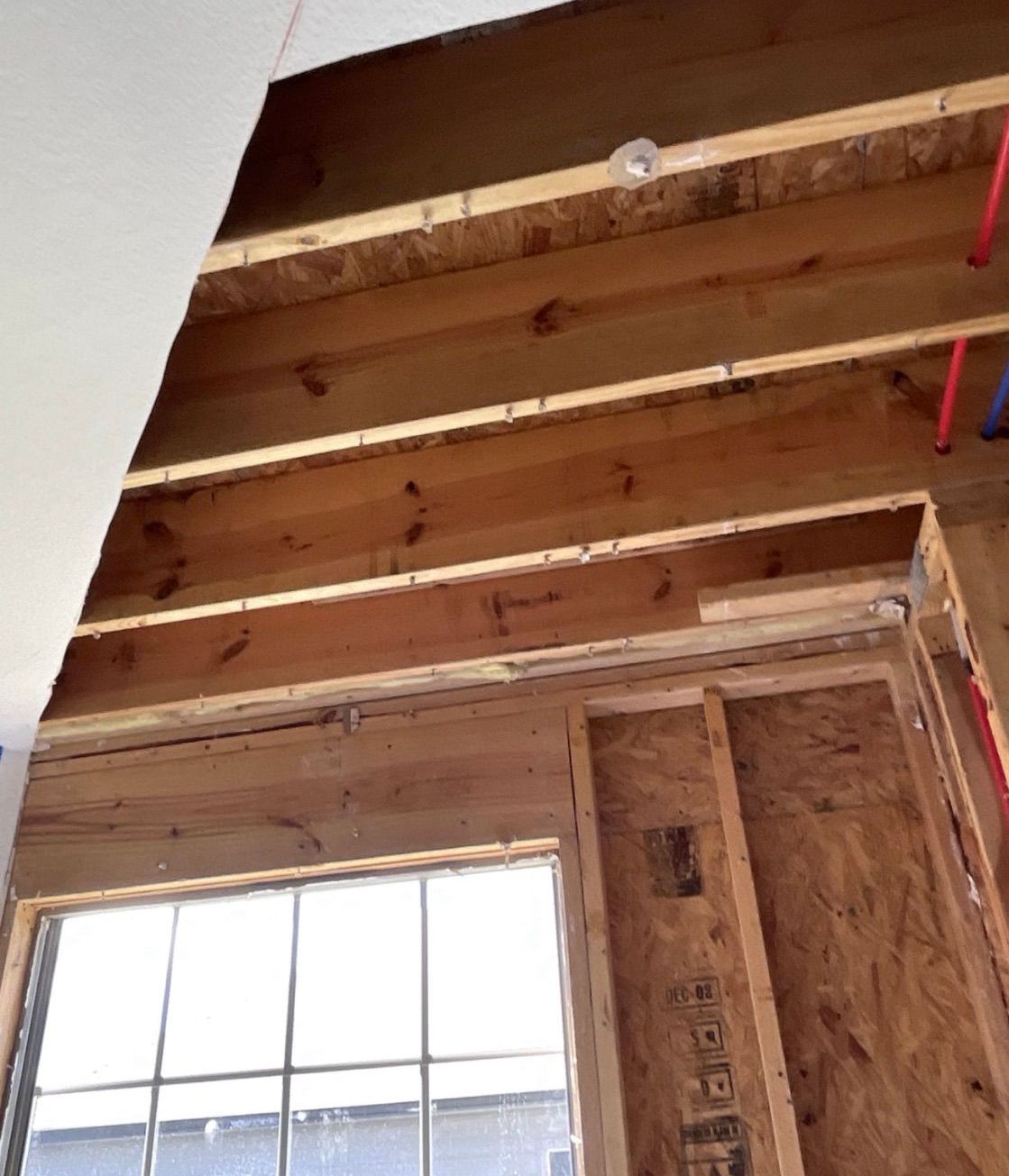 Interior view of a building under construction. Wooden beams and plywood walls surround a window frame. Blue and red pipes are visible.
