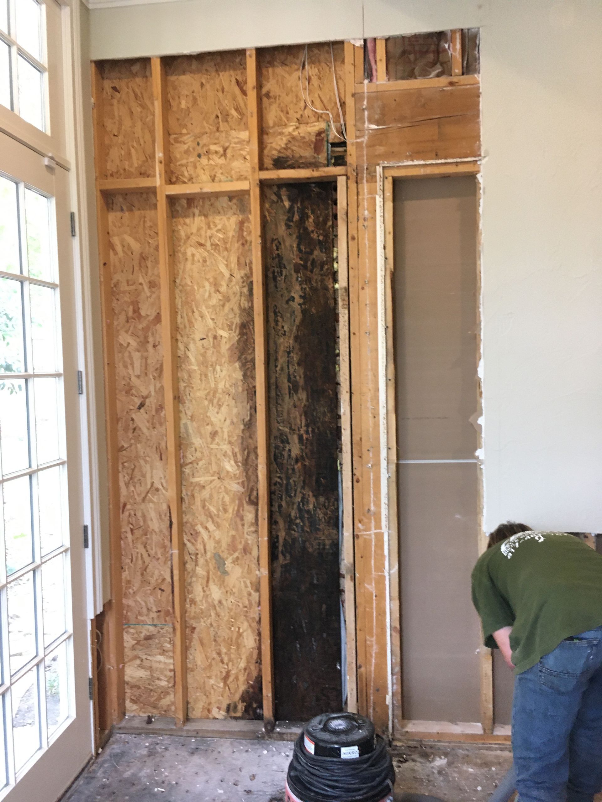 A wall's interior is exposed, revealing dark staining. A person in green is working on the right side of the wall.