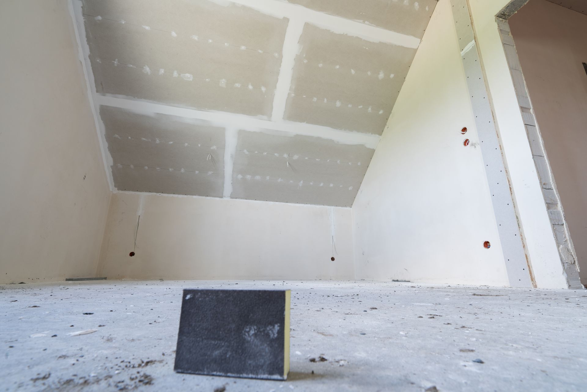 An unfinished room with drywall on the ceiling and walls. A sanding block sits on the floor.