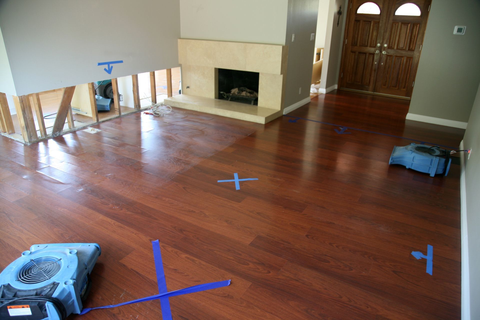 A room with hardwood floors, a fireplace, and fans. Blue tape crosses and exposed wall frame suggest water damage.
