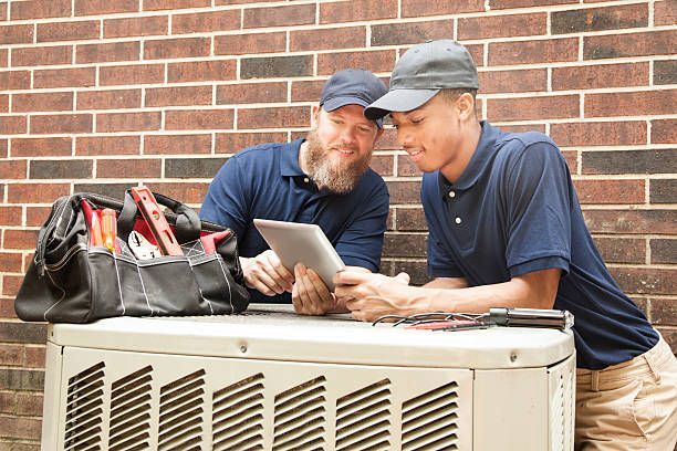 Two HVAC technicians looking at a tablet near an outdoor unit. One has a beard. Brick wall backdrop.