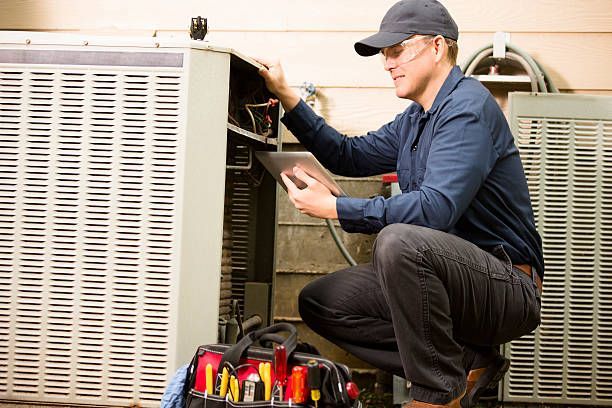 HVAC technician squatting, inspecting an air conditioning unit, holding a tablet, and tool bag.