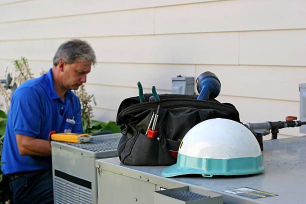 HVAC technician in blue shirt inspecting equipment outside a building with a tool bag and hard hat nearby.