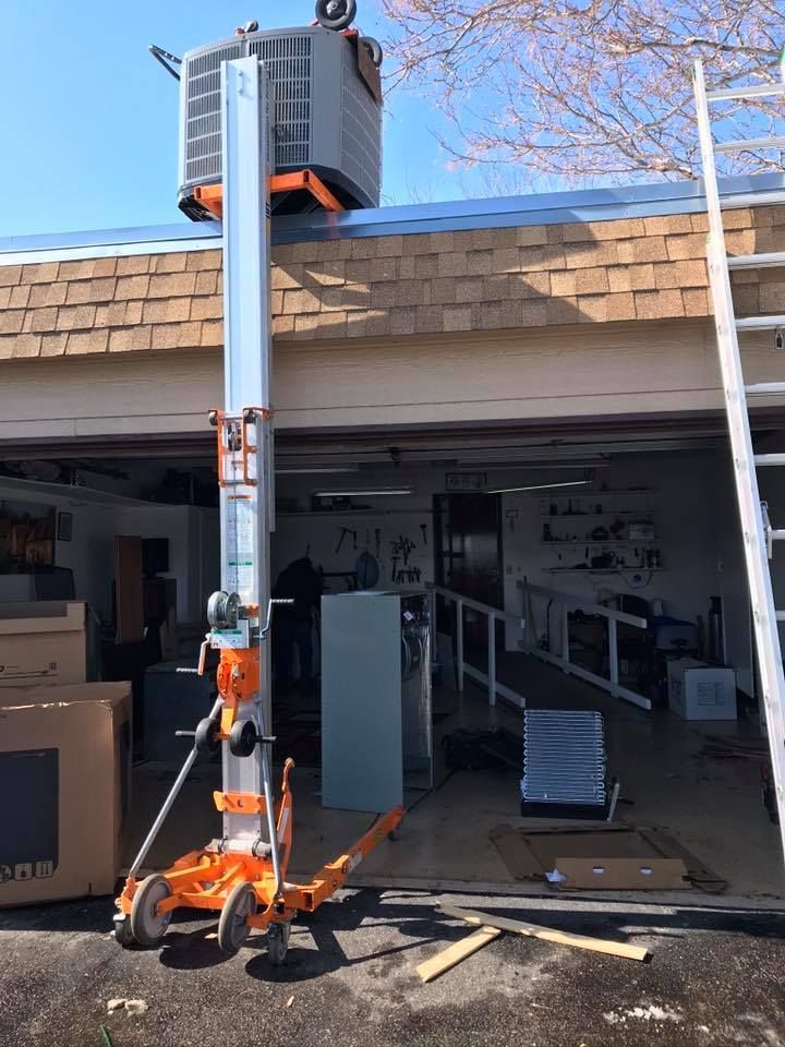 Orange lift raises air conditioner to roof. Garage background, ladder nearby. Sunny day.