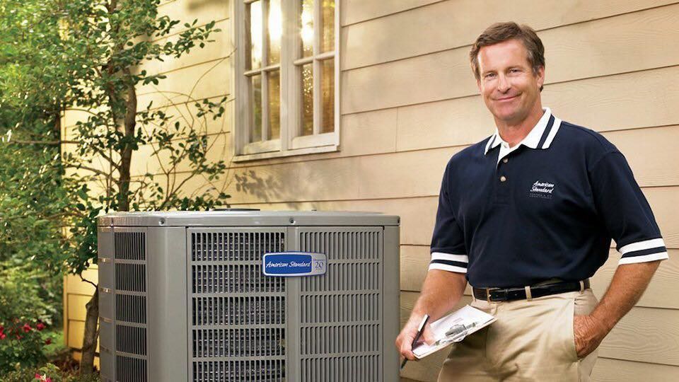 HVAC technician smiling next to an air conditioning unit outside a house.