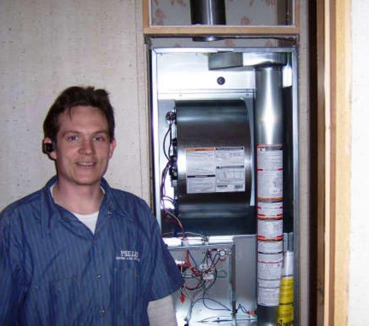 HVAC technician in blue shirt stands by furnace, smiling.