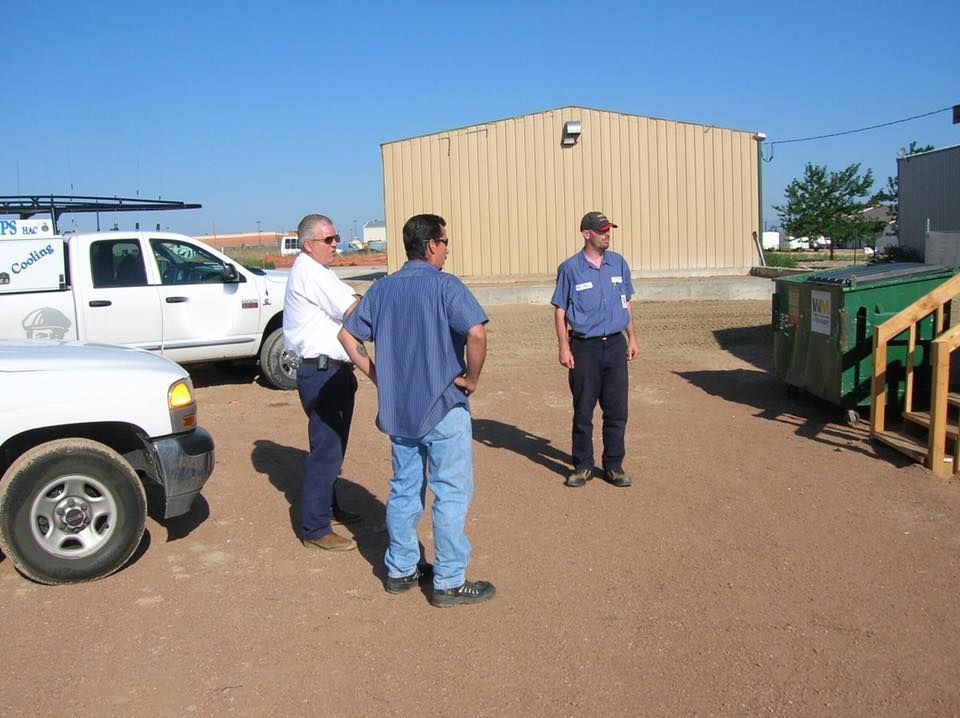Three men in a gravel lot near buildings and trucks; one wears a blue uniform.