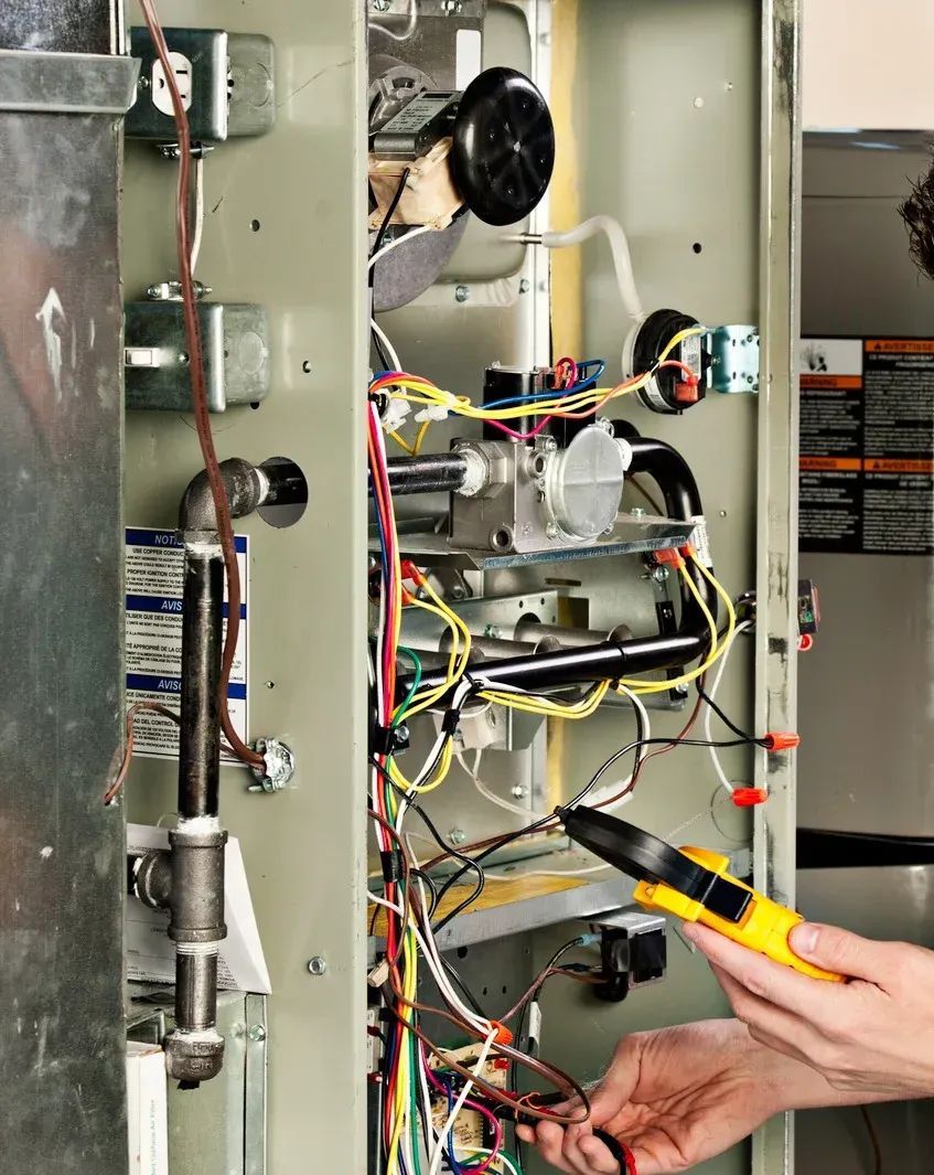 Person using a multimeter to inspect wiring inside a furnace cabinet, assessing electrical components.
