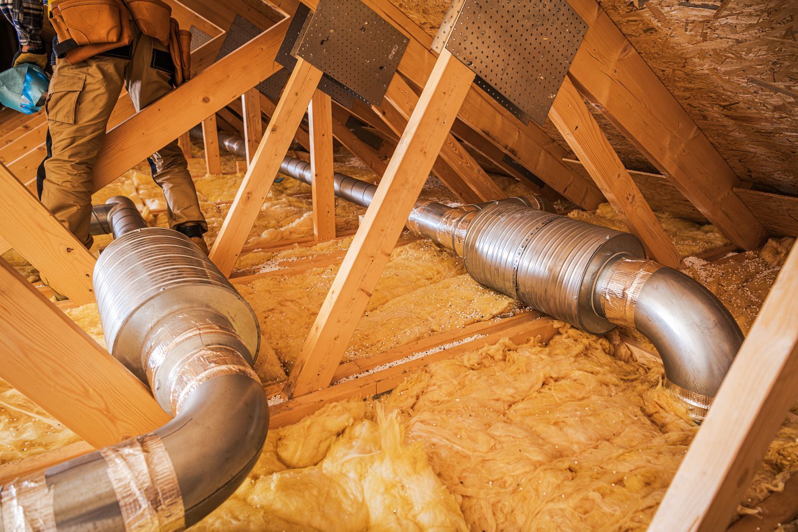 Person in an attic installing HVAC ductwork among wooden beams and insulation.