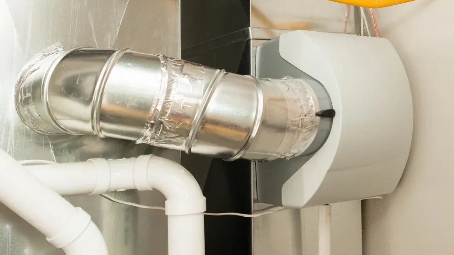 A humidifier attached to silver ductwork and white plumbing, in a home setting.
