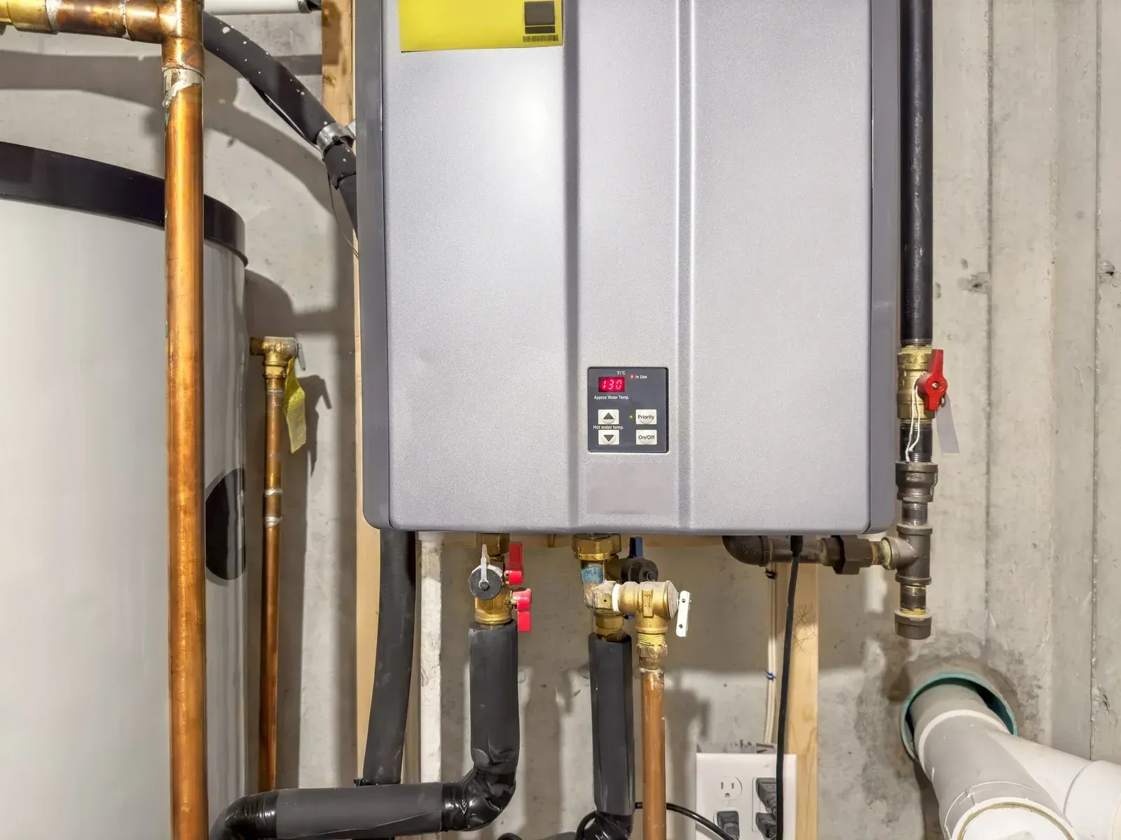 Tankless water heater and hot water tank on a basement wall with copper and black pipes.