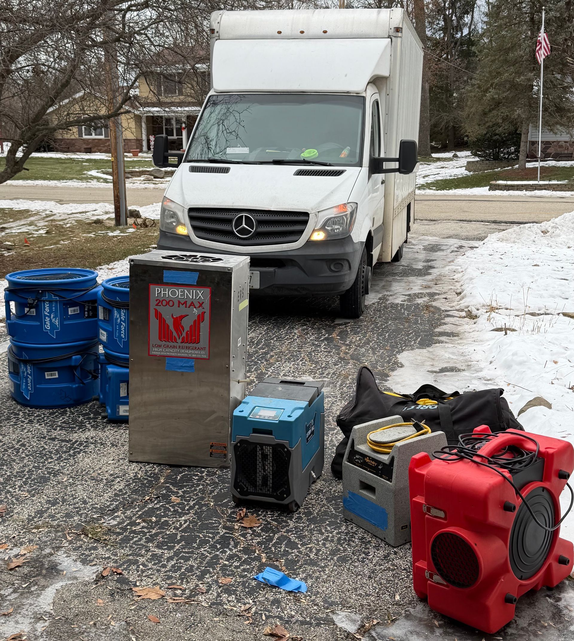 A white van parked outdoors with restoration equipment, including blue bins, a metal unit, and air movers, on the ground.