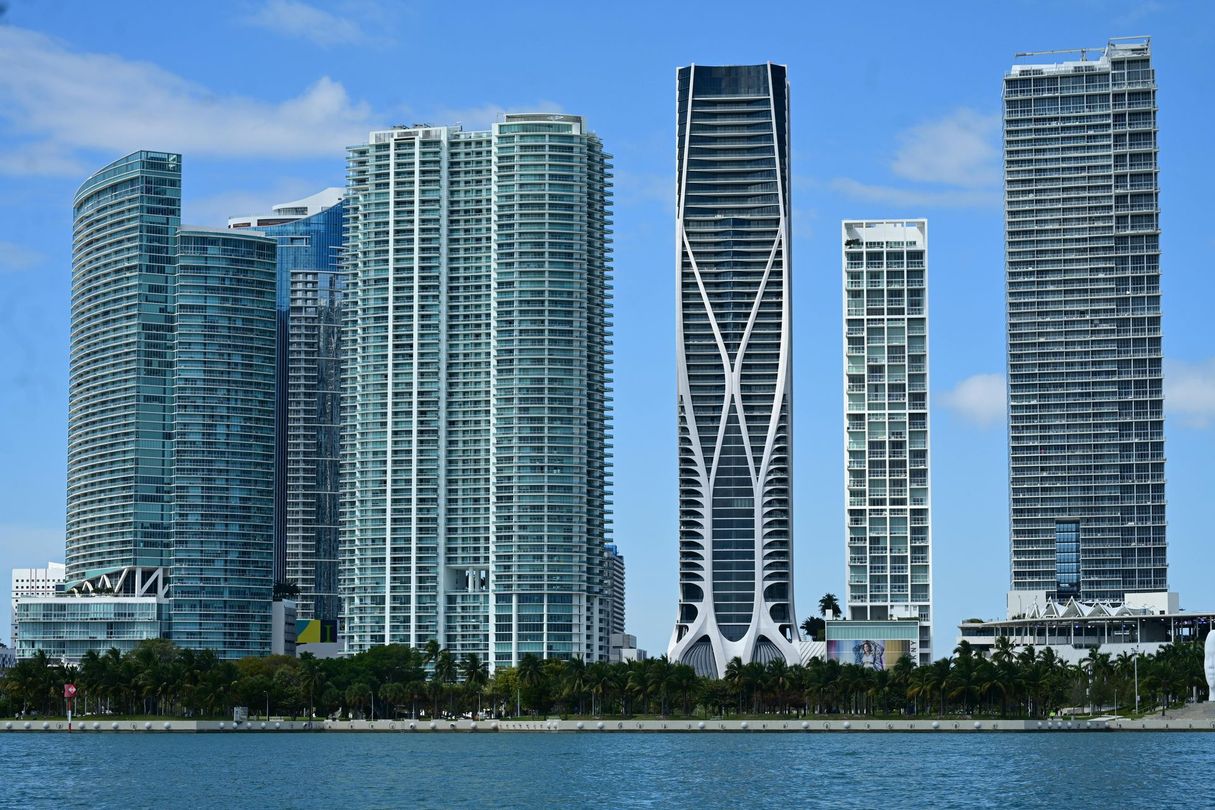 Miami skyline with modern skyscrapers against a blue sky, trees in the foreground, and water below.