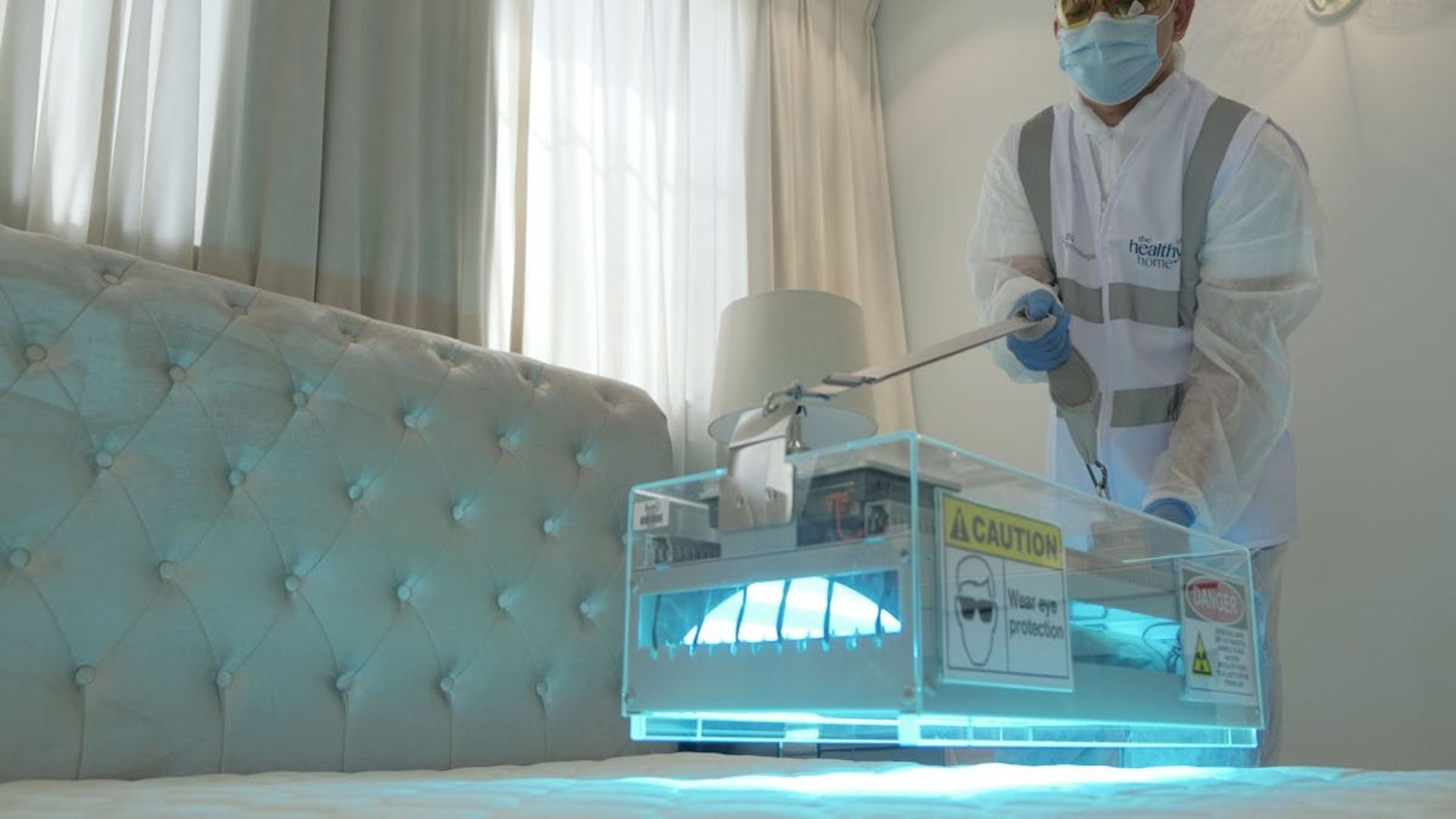Una persona con equipo de protección utiliza un dispositivo de desinfección UV en una cama en una habitación blanca.