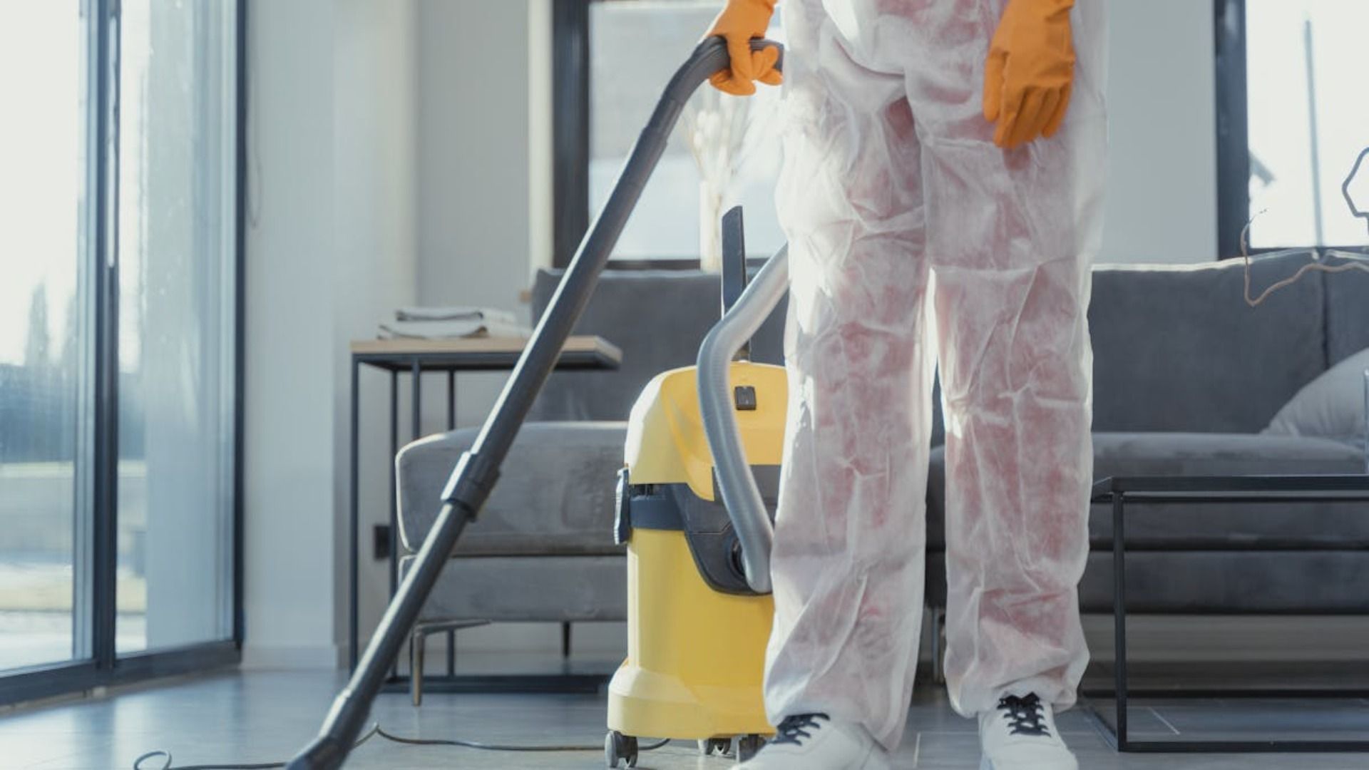 Una persona con traje protector blanco aspira una sala de estar, lleva guantes naranjas y utiliza una aspiradora amarilla.