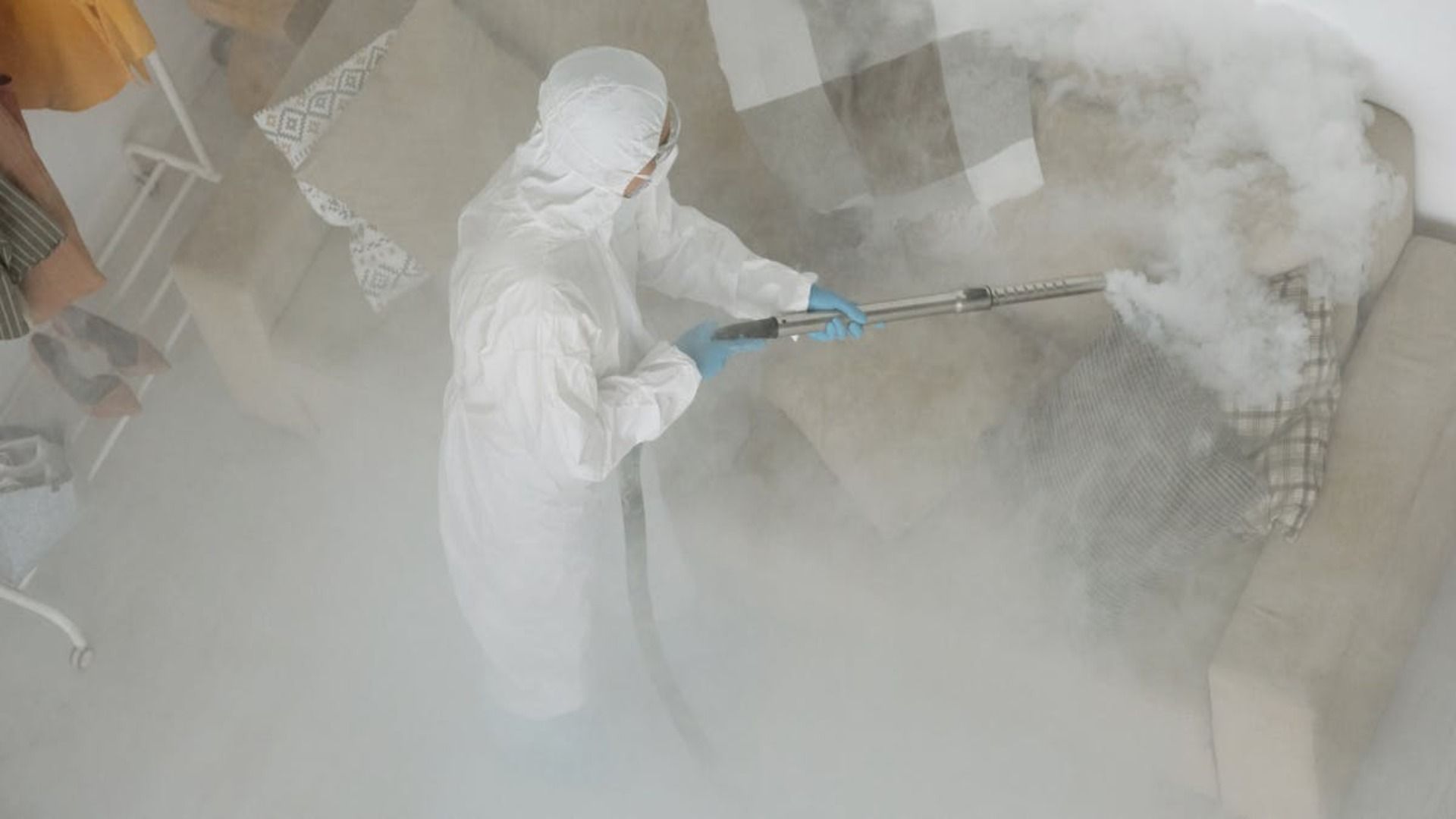 Persona con traje de materiales peligrosos desinfectando un sofá con una máquina de niebla en una sala de estar.