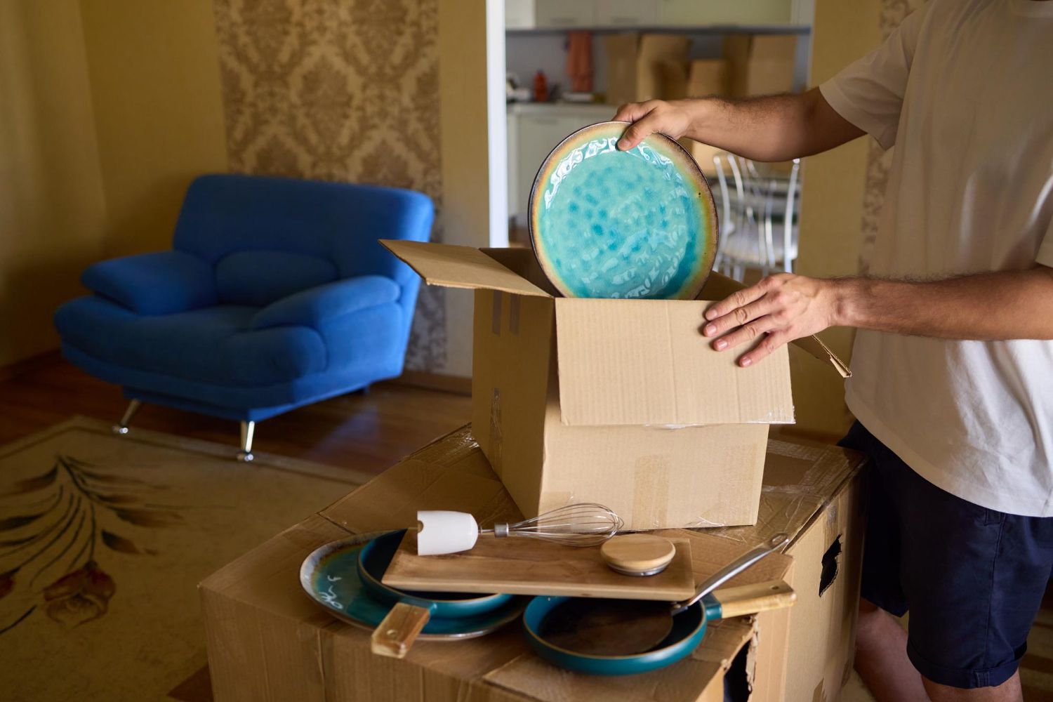 Man packing dishes in a cardboard box in a room with a blue armchair. He holds a teal plate.