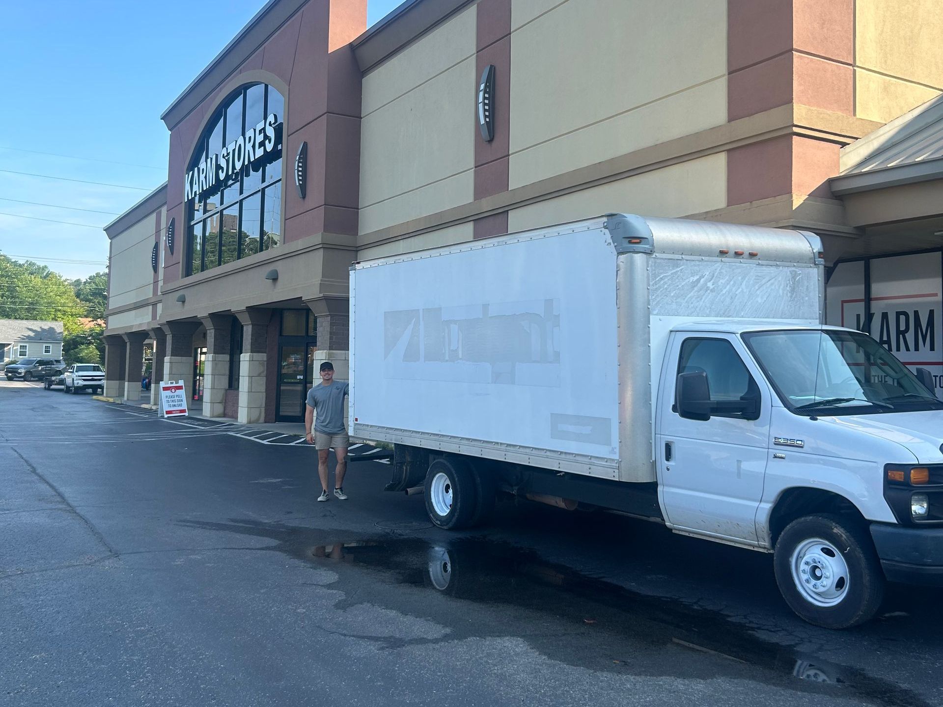 A man stands near a white box truck parked in front of a beige and brown store. The sky is blue.