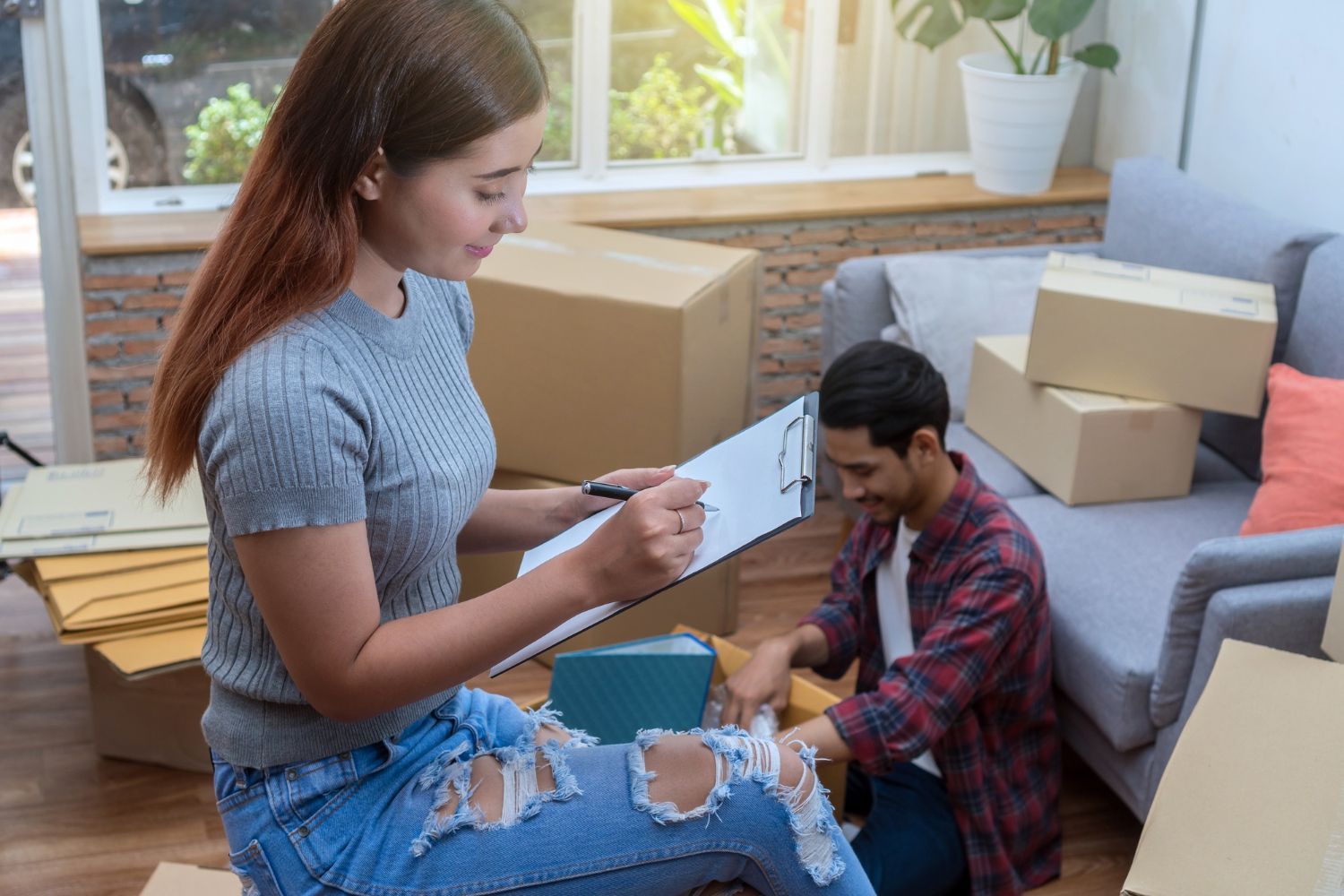 Woman with clipboard and man packing boxes in a living room; sunlight streams in.