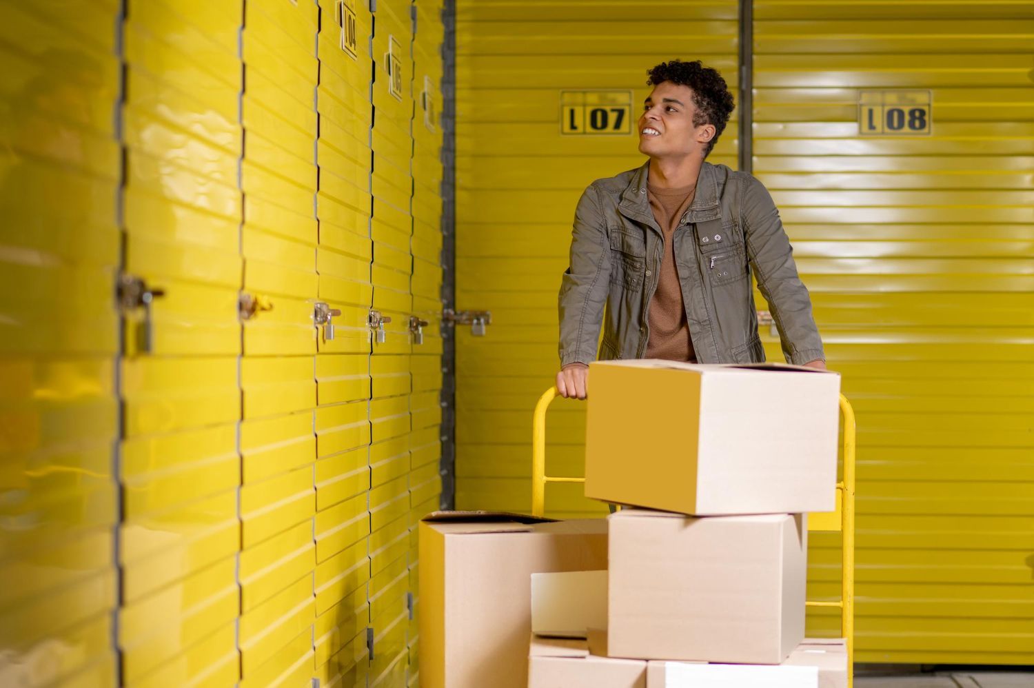 A person standing in a yellow storage unit, leaning on a cart with cardboard boxes, looking upward.