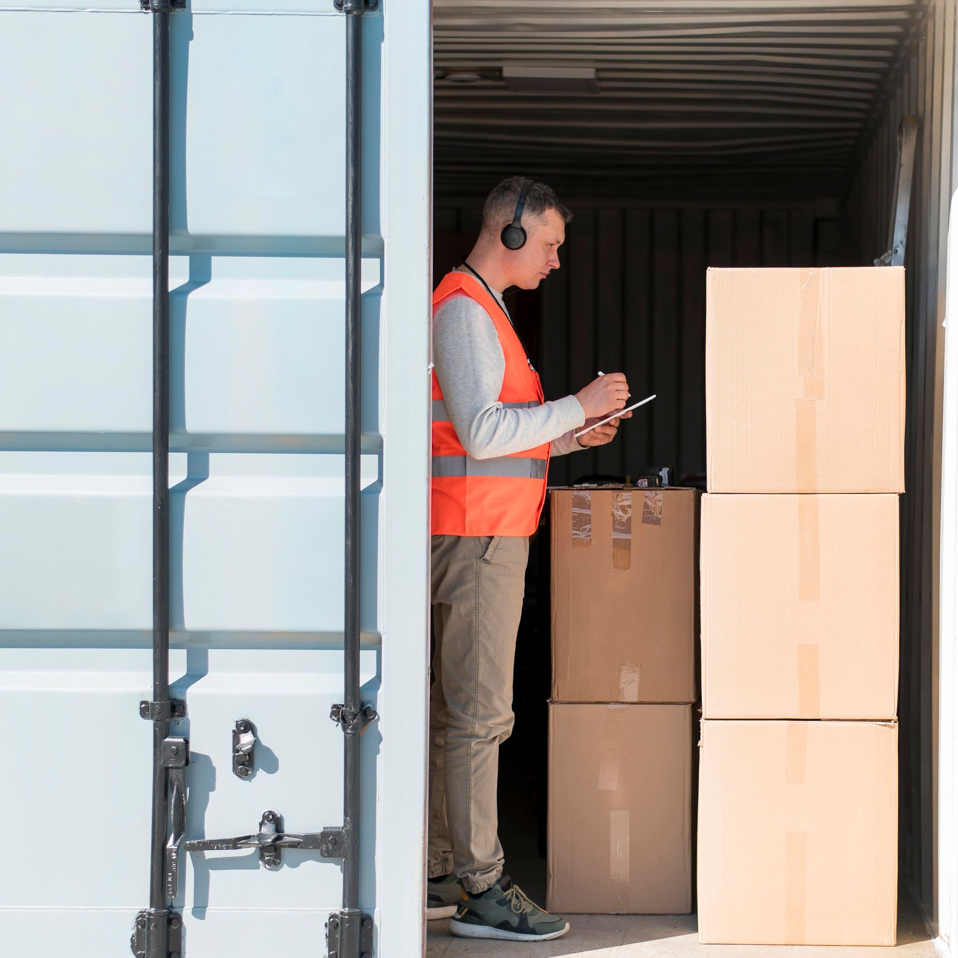 A man wearing headphones is standing in a shipping container looking at a tablet.