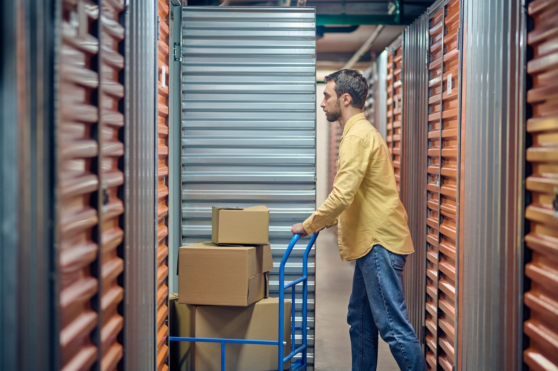 Man in yellow shirt and jeans pushes a hand truck with cardboard boxes down an aisle of storage units.