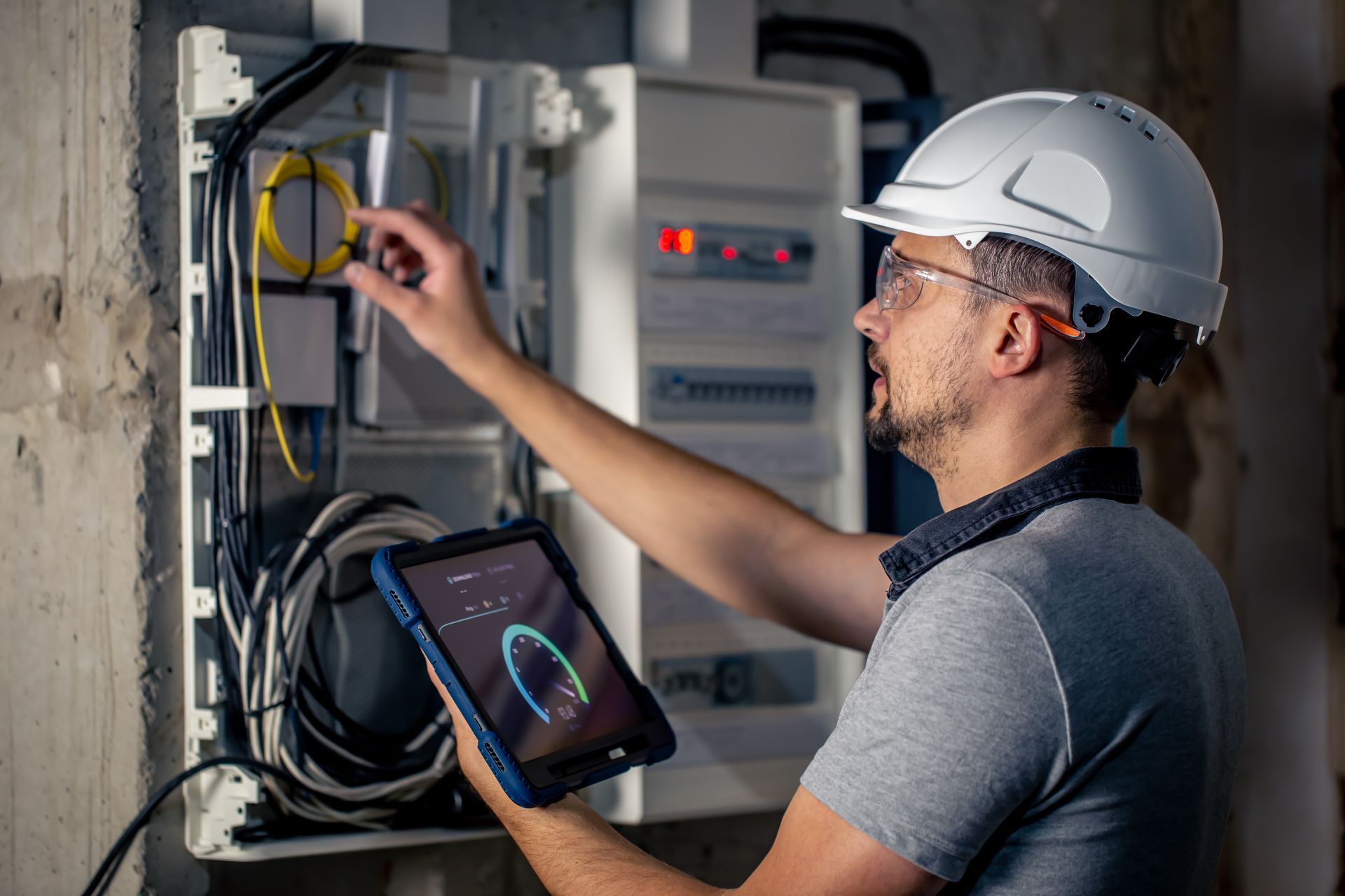 A man in a hard hat is using a tablet to look at an electrical box.