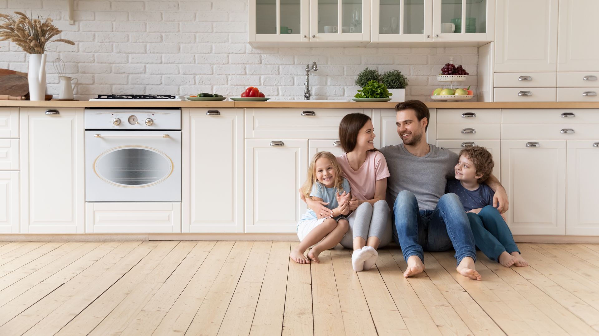 A happy family of four sits on the floor of their bright, modern kitchen, smiling and looking at each other.