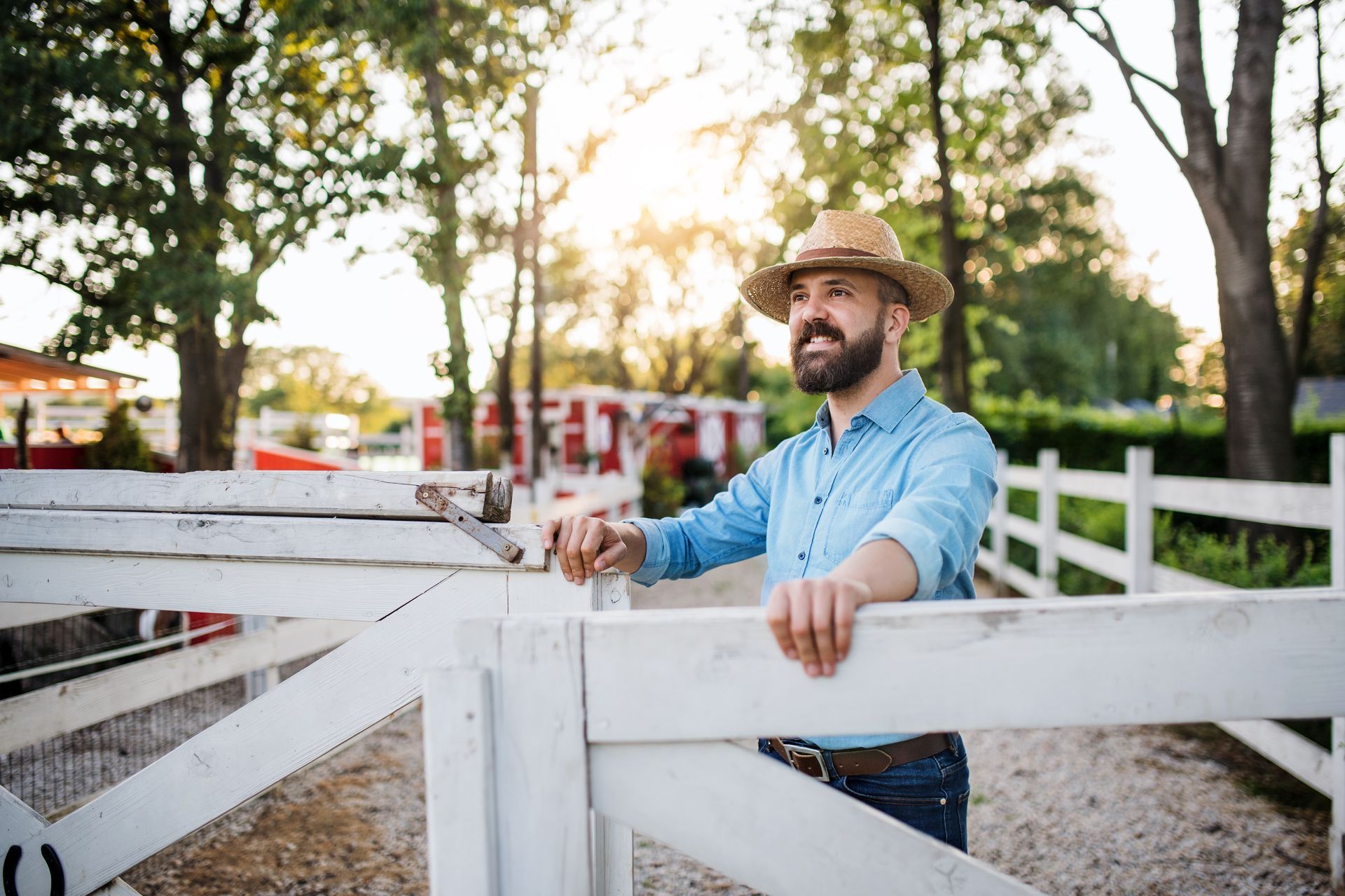 Man in straw hat smiles, leaning on white fence in a sunny outdoor setting.