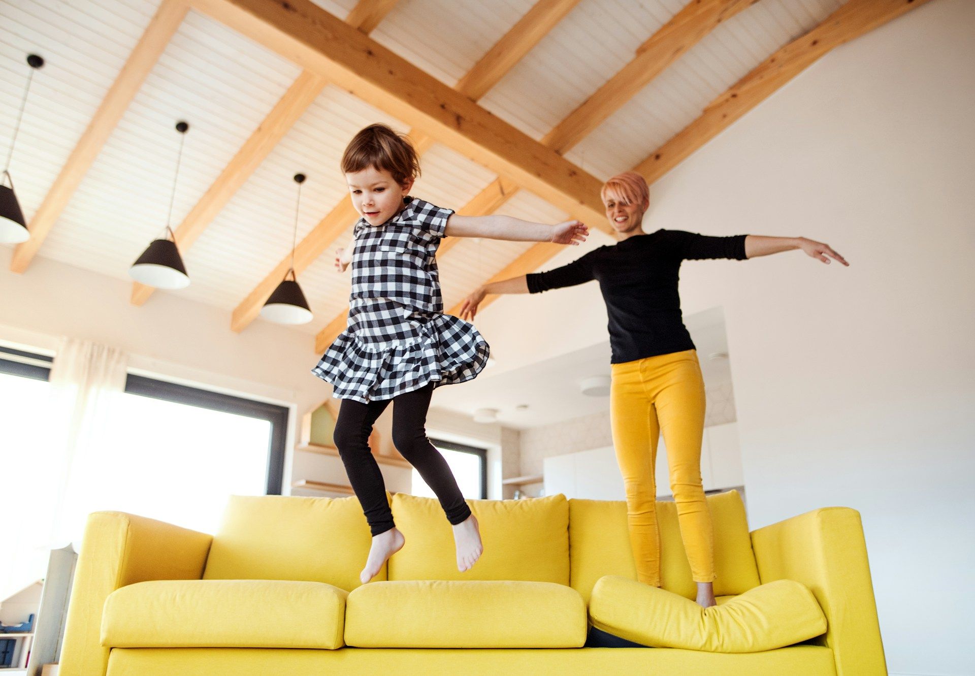 Woman and child jumping joyfully on a yellow couch in a bright living room.