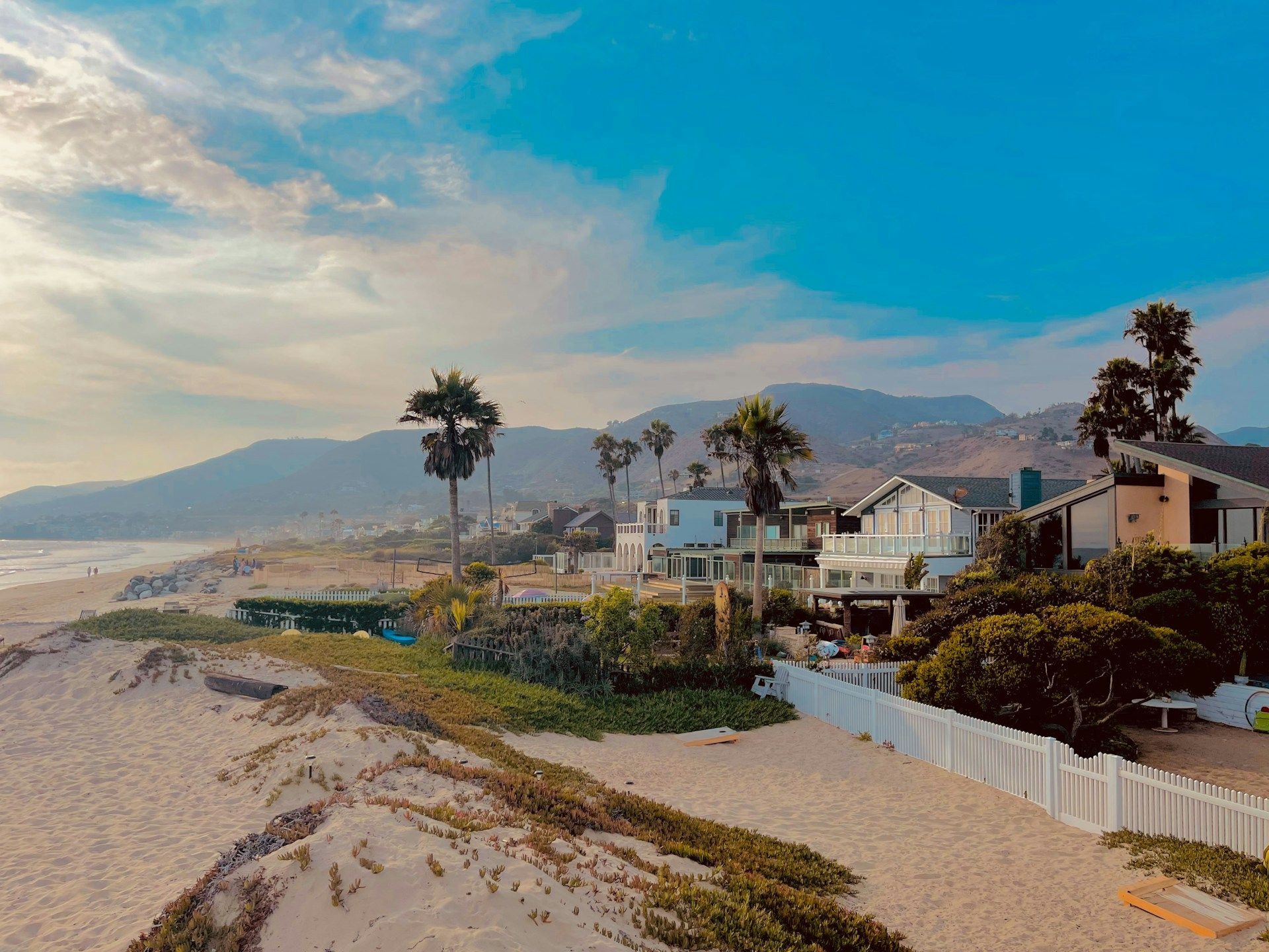 Beachfront homes under a blue sky with palm trees and mountains in the background.
