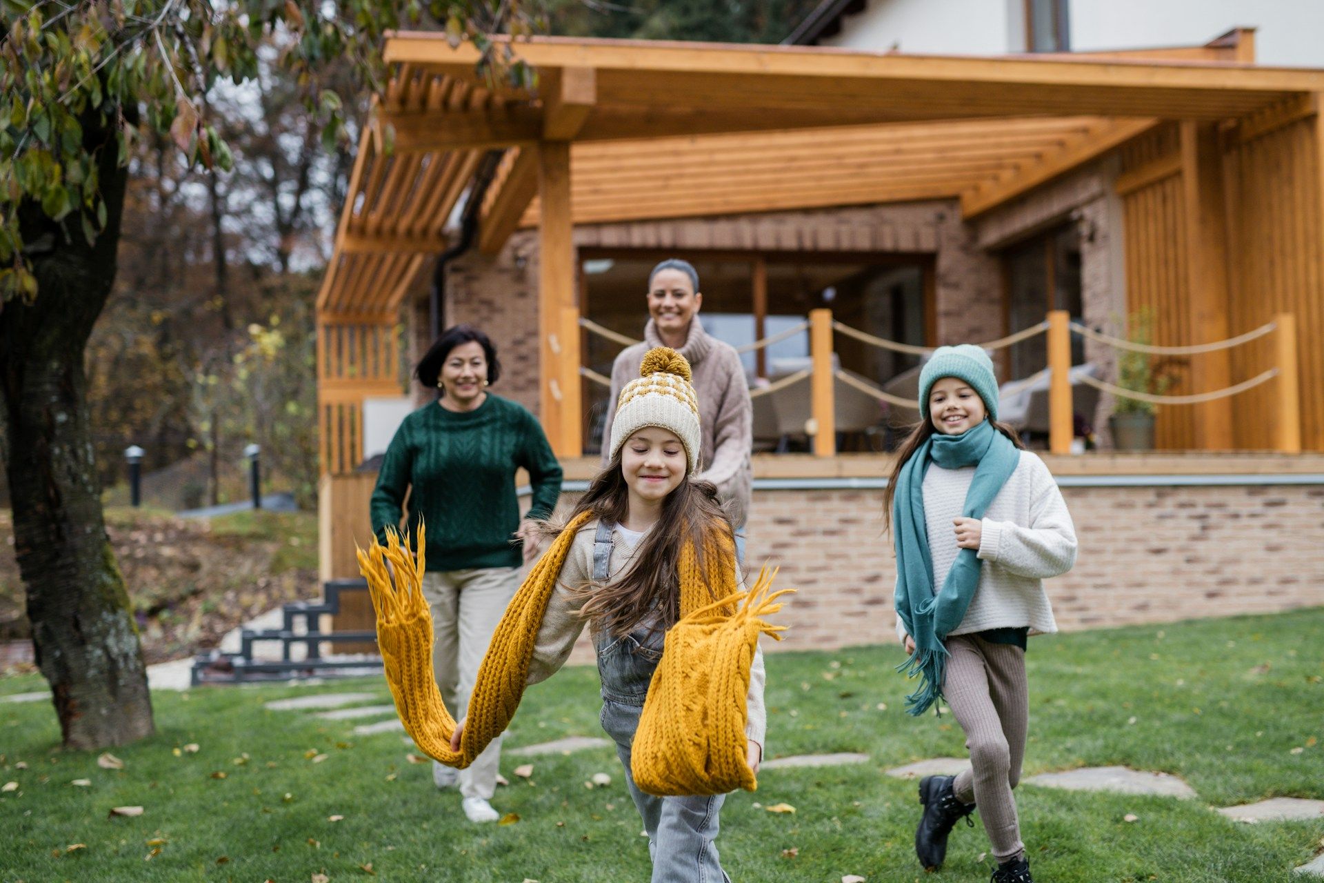 Four people, including two children, running in a grassy yard near a house, wearing winter hats and scarves.