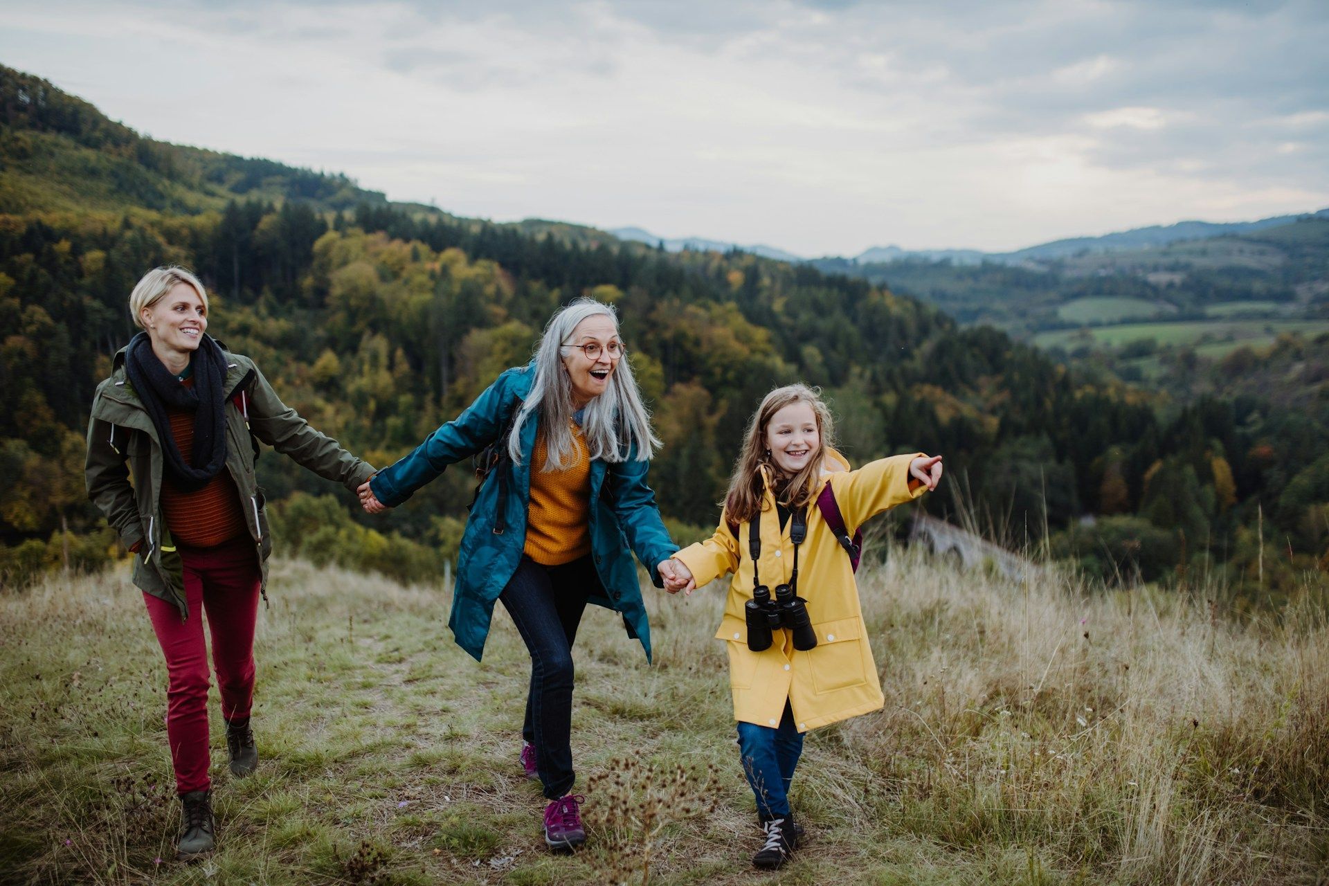Three people, including a child, hold hands hiking on a hillside with a forest backdrop.