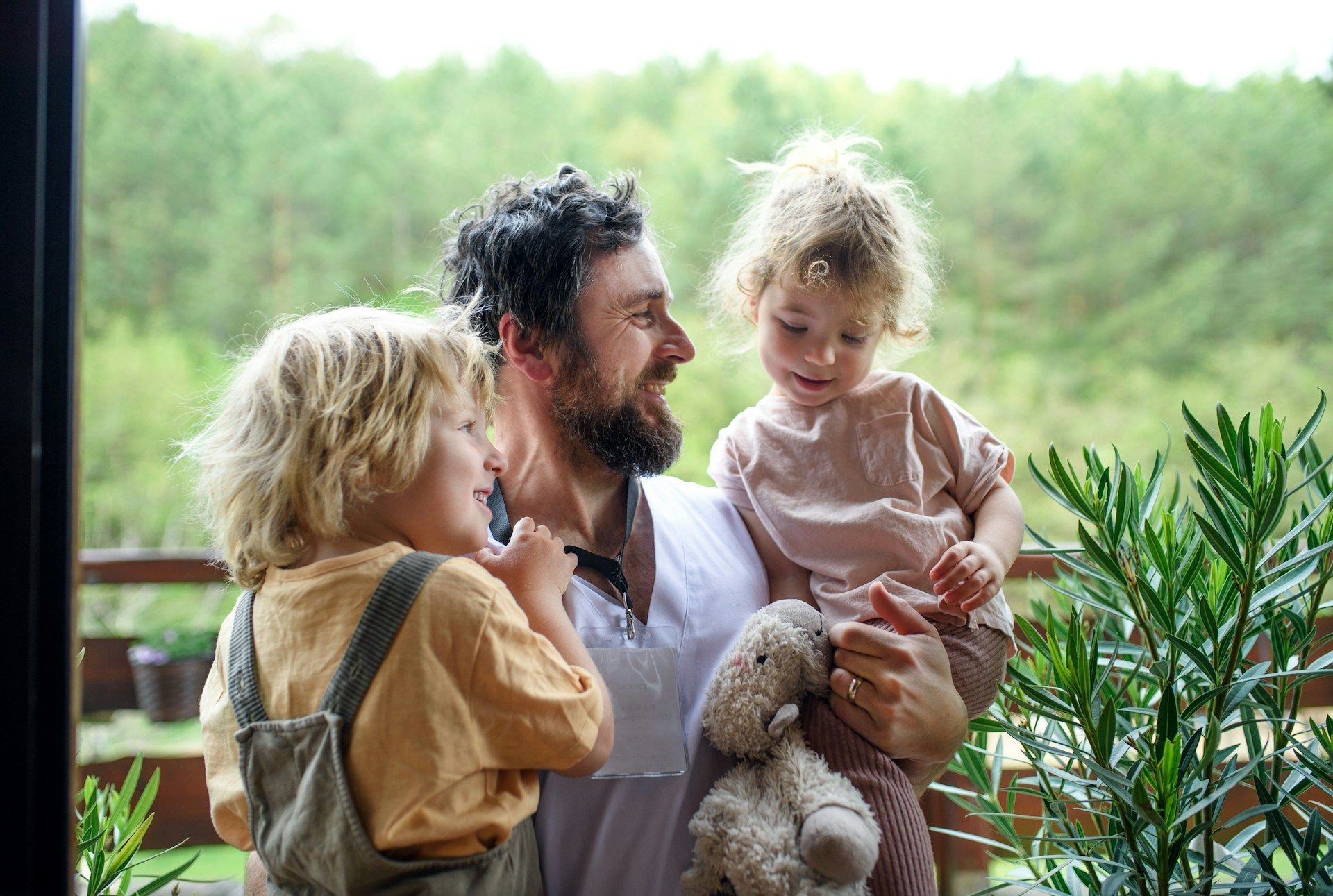 Father holds two young children on a balcony, smiling. Blonde hair, trees in background.