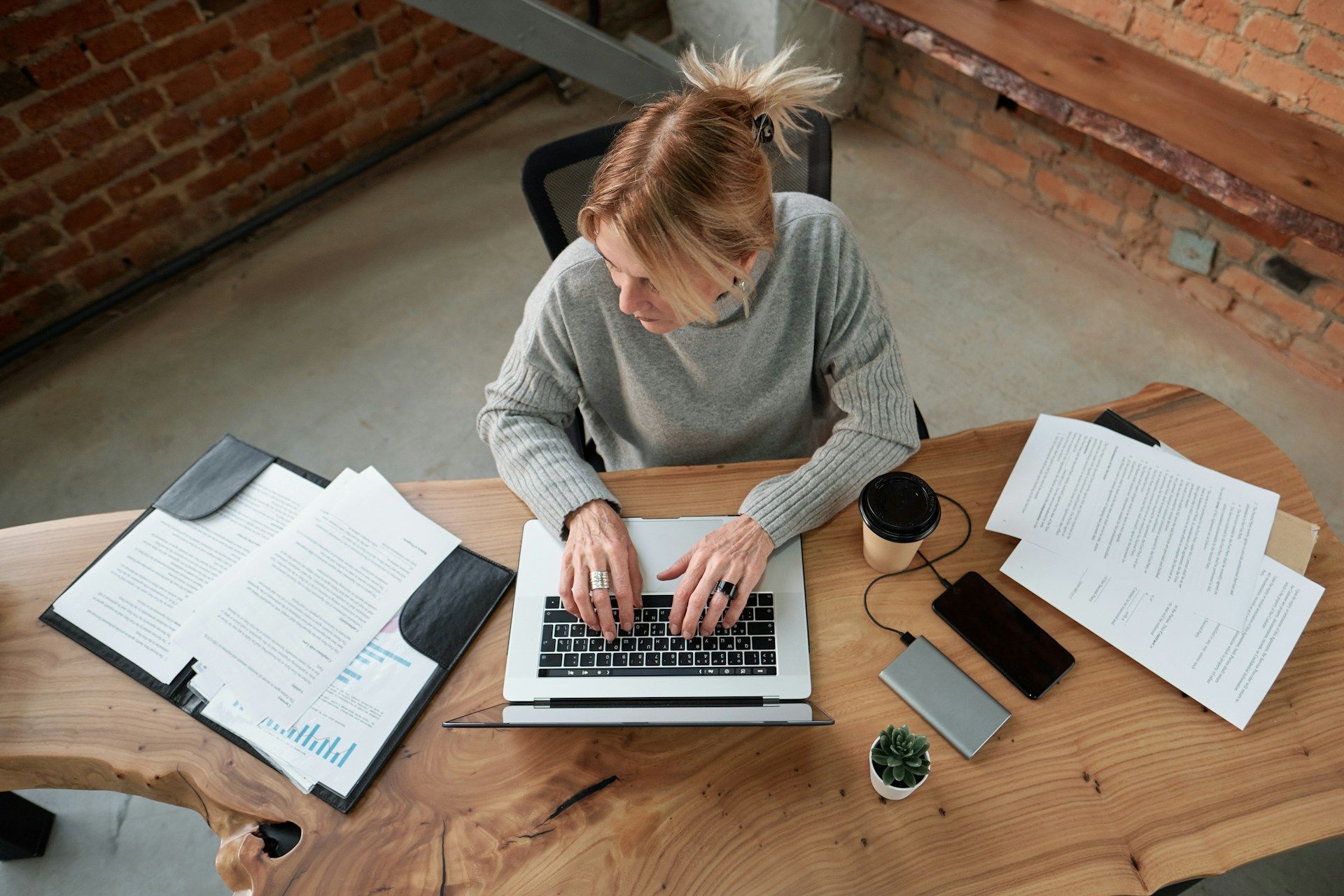 Woman typing on laptop at wooden desk, papers and coffee present.