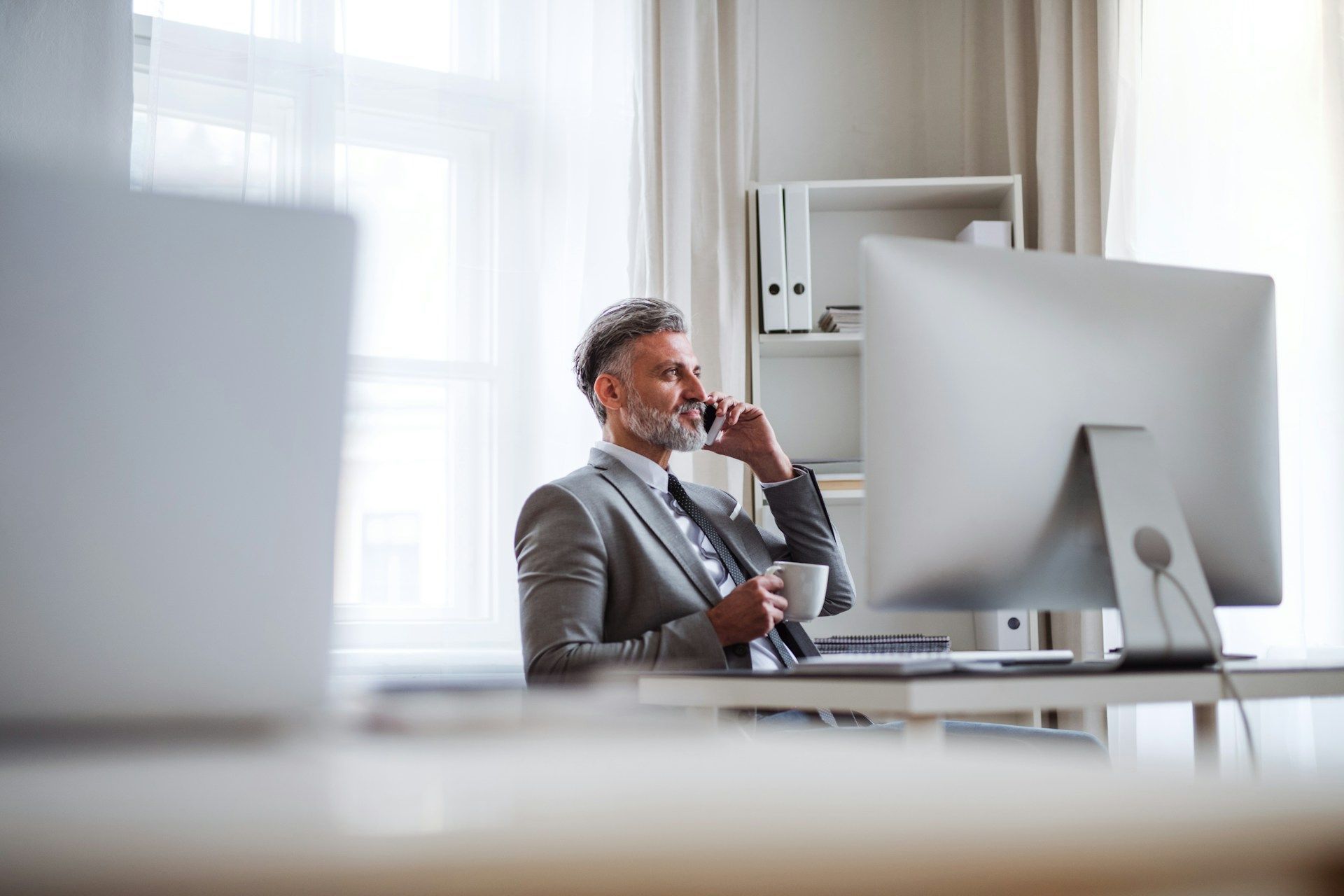 Man in suit on phone, sitting at desk with computer and mug in a bright office.