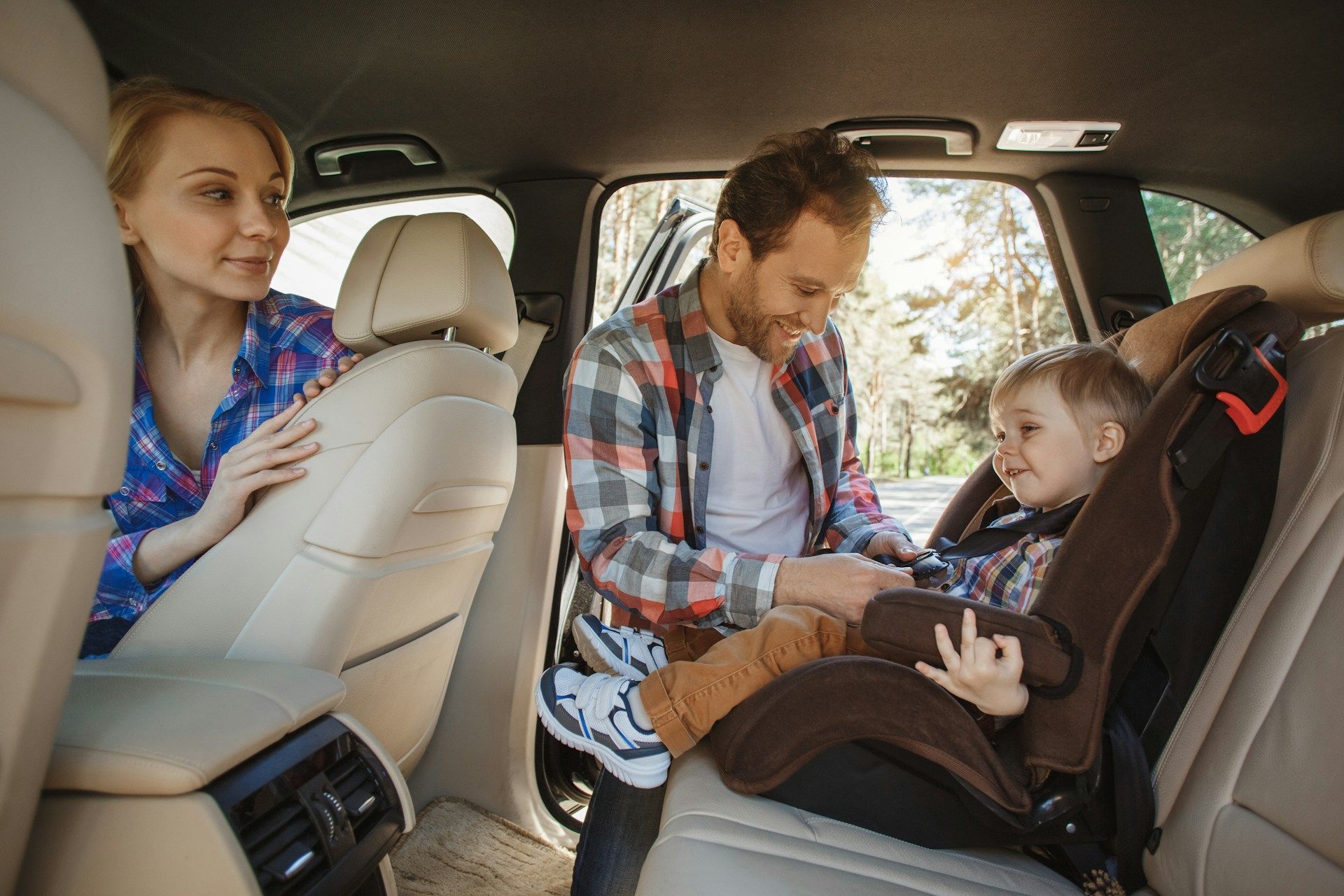 Parents securing a child in a car seat inside a vehicle; sunny outdoors in background.