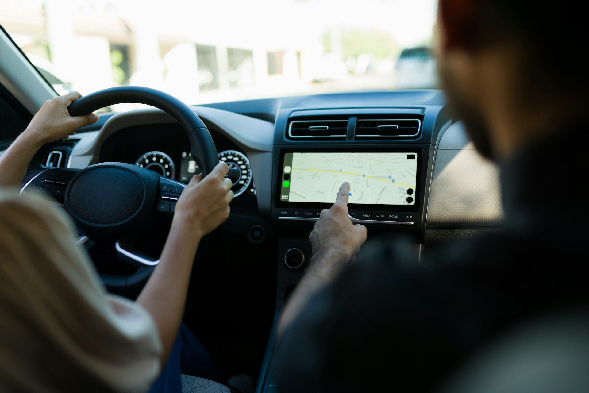 Inside a car: Person driving, other person pointing at a navigation screen, guiding them.
