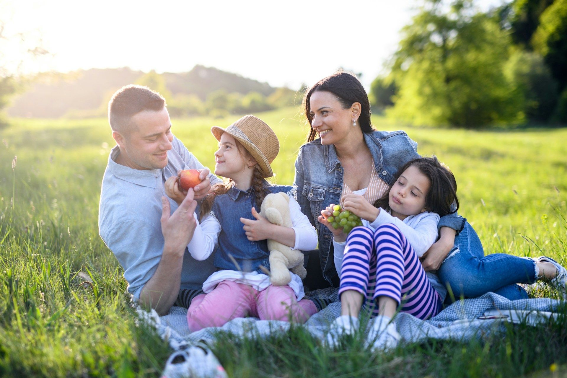 Family of four picnicking in a sunny field, smiling and enjoying fruit.