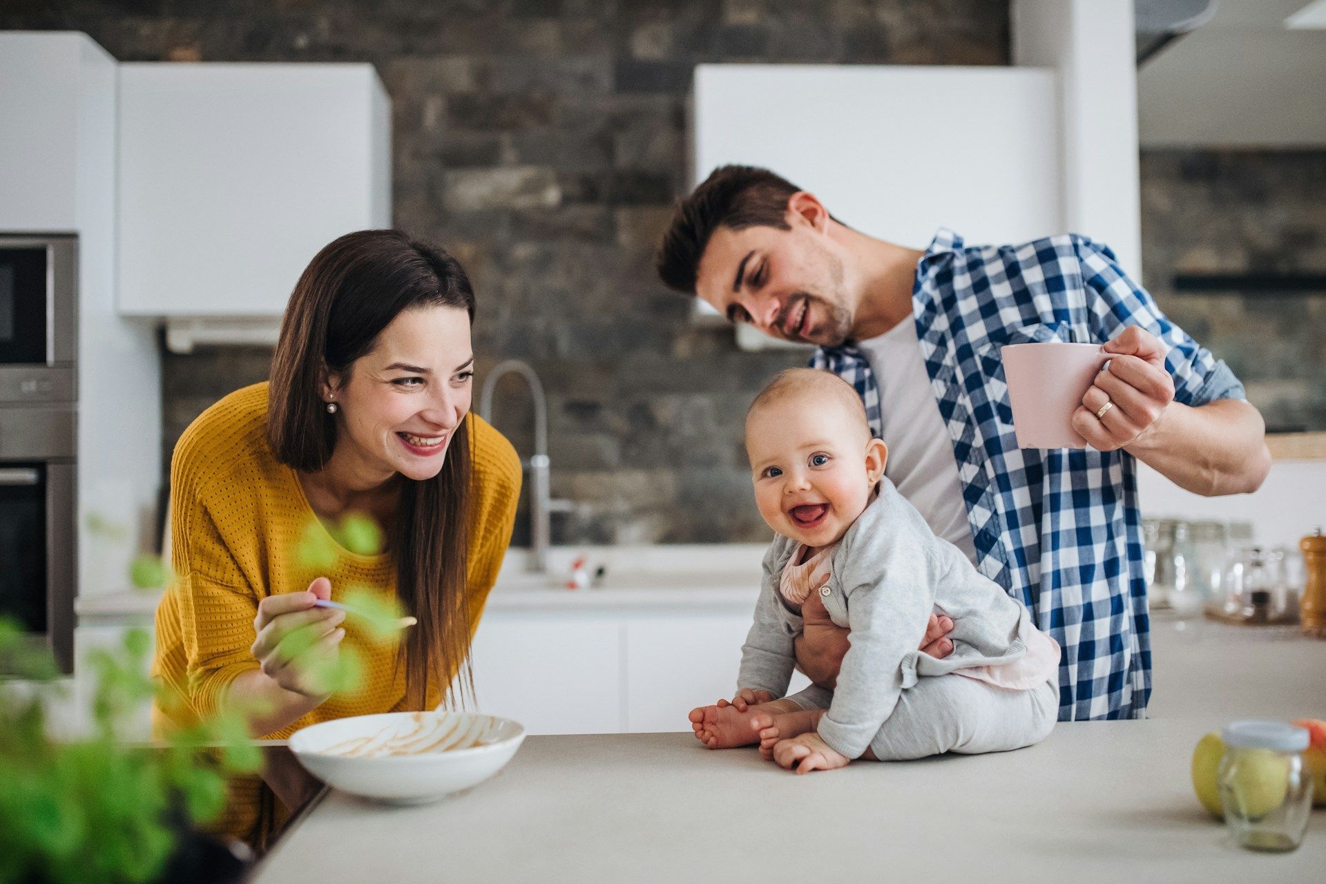 Family in a modern kitchen: baby on counter laughs, mom smiles, dad holds coffee cup, background is white.