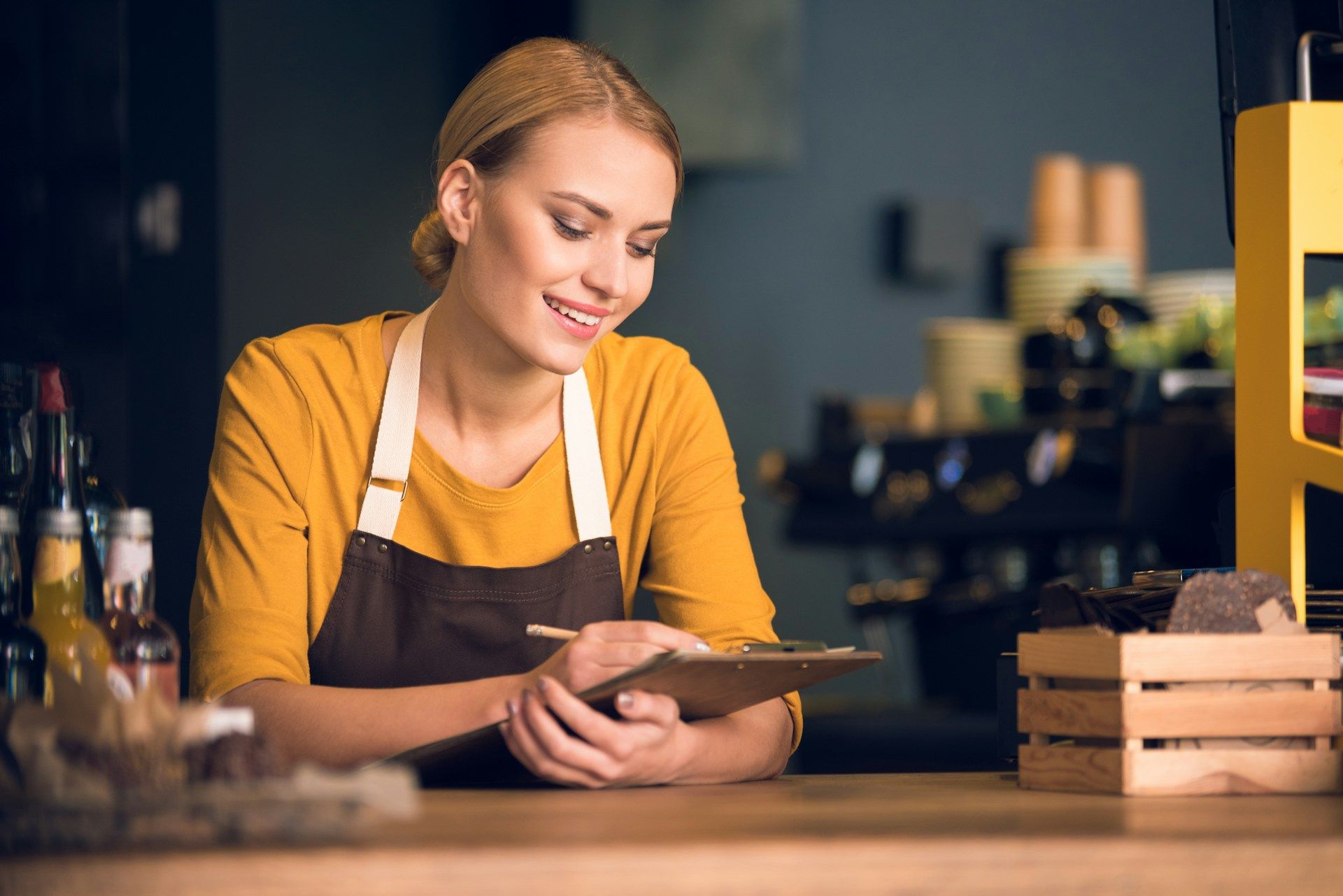 Woman in apron smiling, taking notes behind a cafe counter.