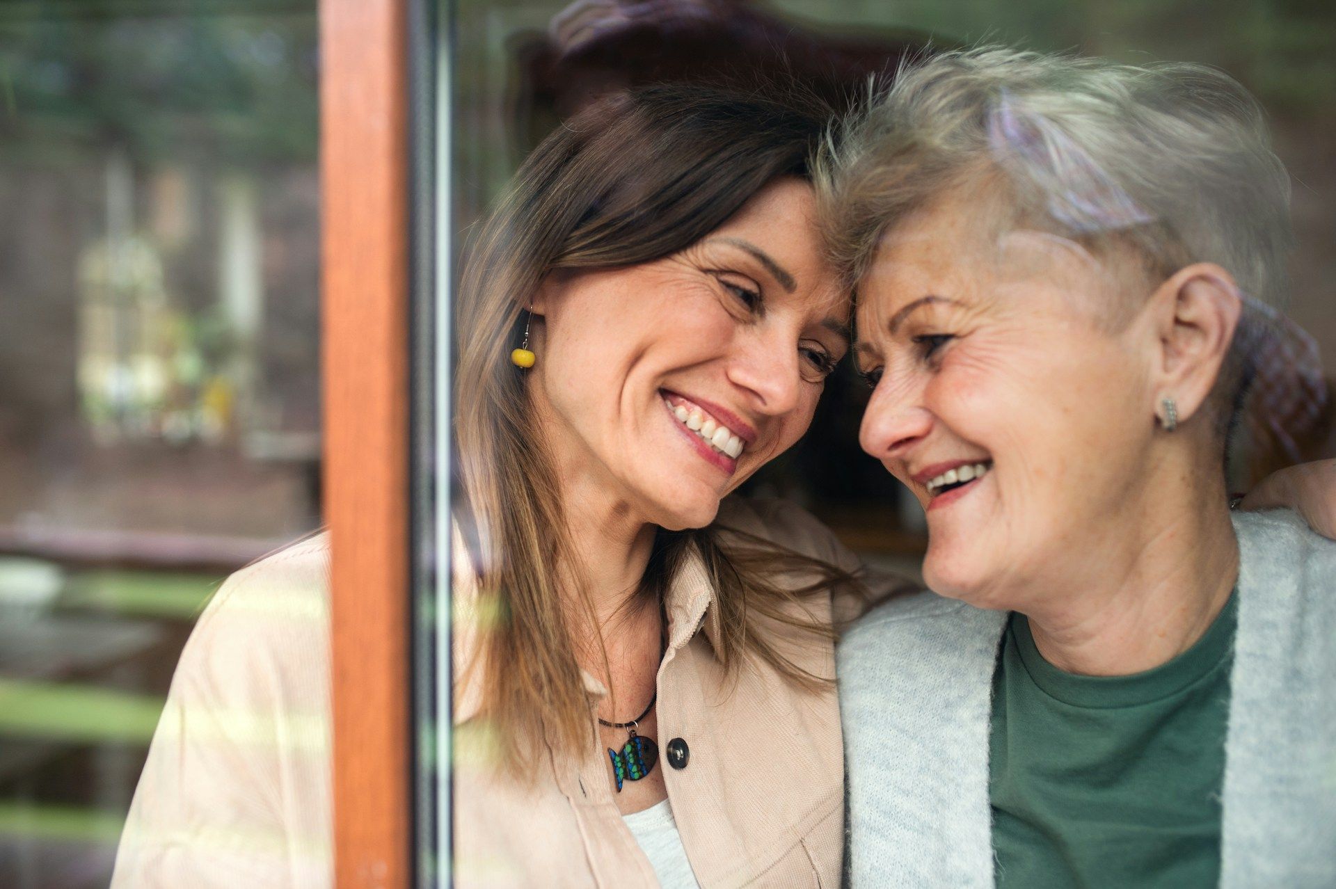 Woman smiles at elderly woman, both touching heads, looking at each other, inside near window.